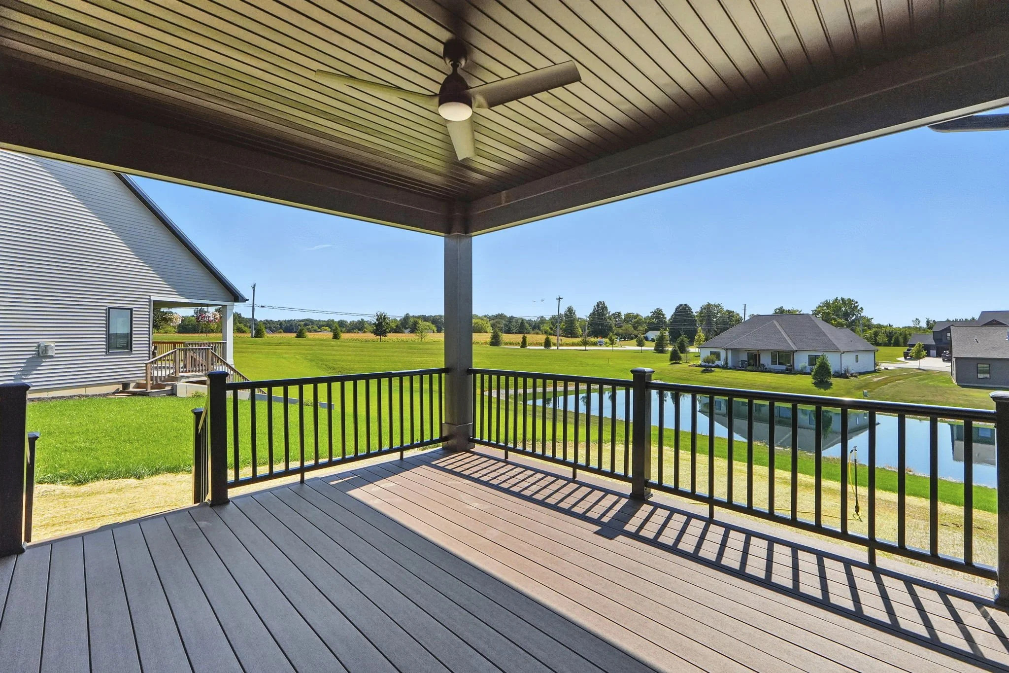 View from a covered porch with a ceiling fan overlooking a grassy yard, a lake, and neighboring houses under a clear blue sky.