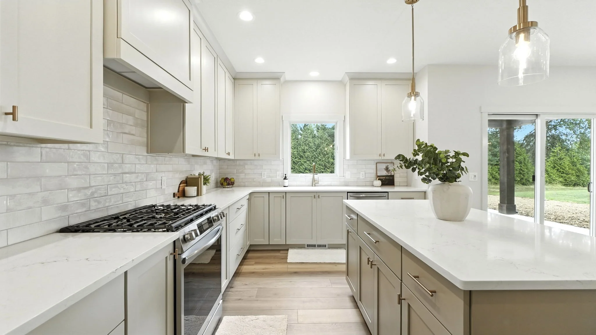 Bright modern kitchen with white cabinets, marble countertops, a window above the sink, and pendant lights over the island. Sliding glass door reveals outdoor greenery.