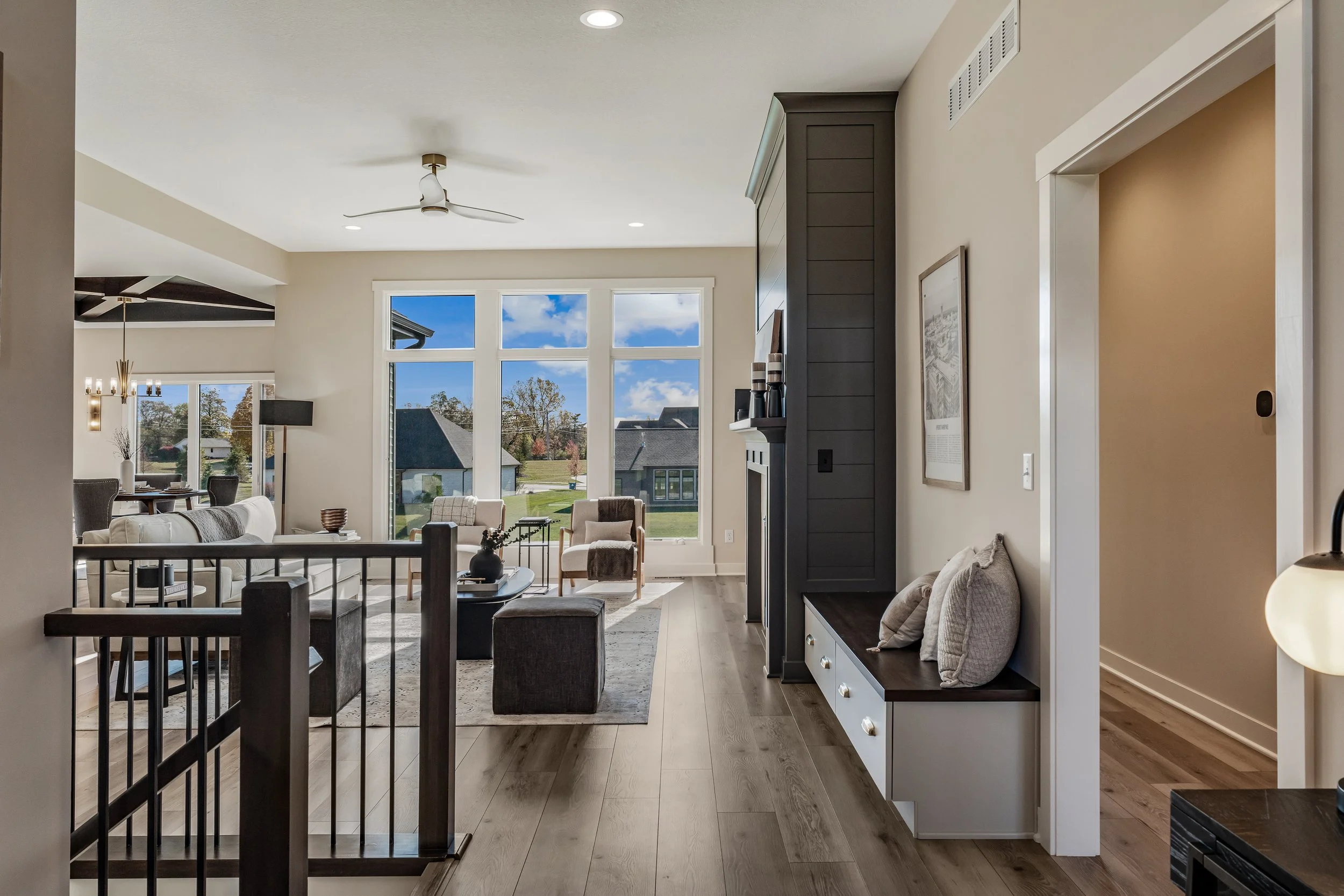 Interior view of a modern living room with large windows, allowing natural light, beige walls, a dark wood accent wall with a fireplace, seating area with chairs and a sofa, and a view of neighboring houses and green lawn outside.