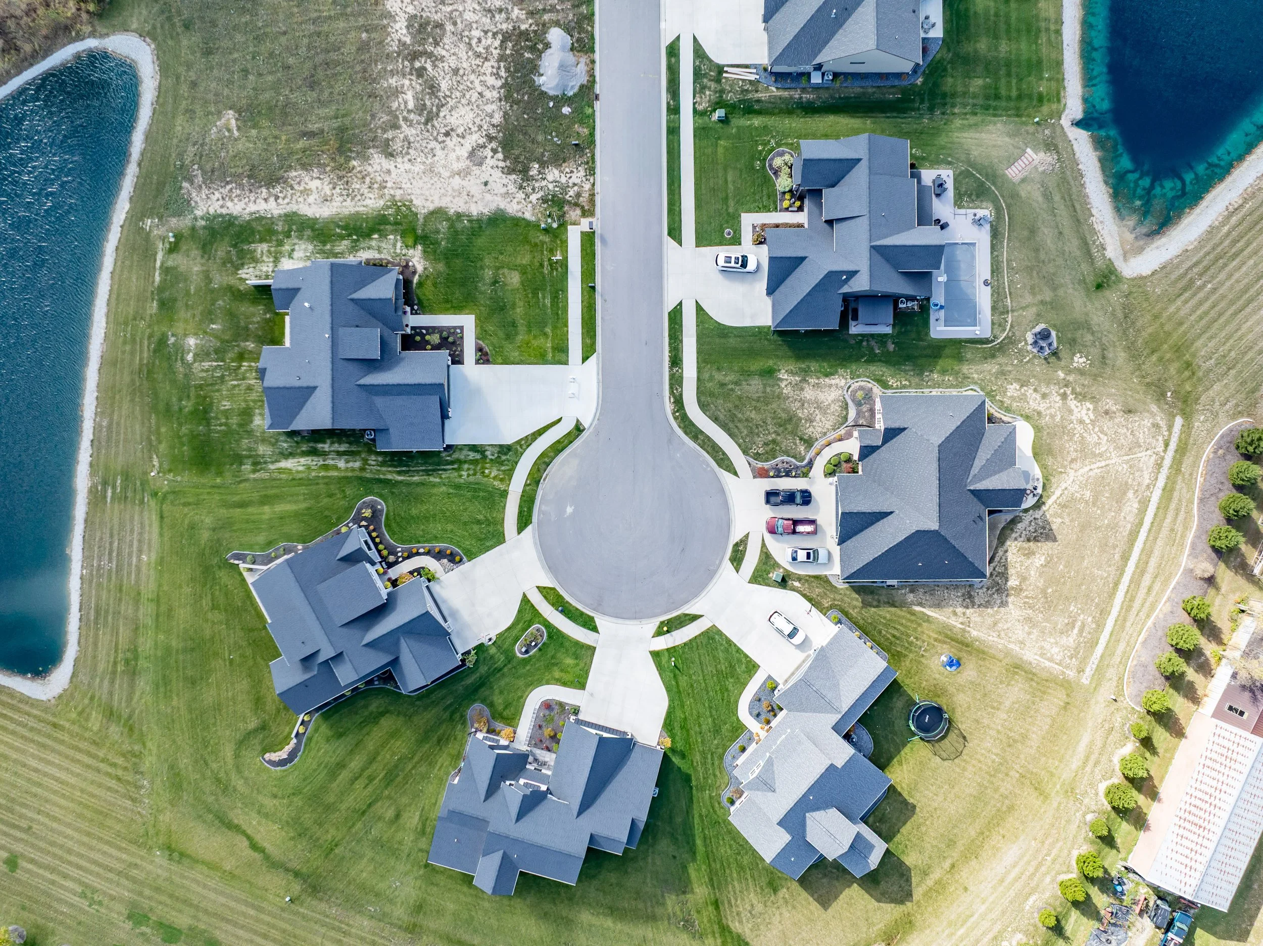 An aerial view of a residential neighborhood showing five houses with driveways, parked cars, landscaped yards, and a roundabout in the center. There are bodies of water and grassy areas surrounding the homes.