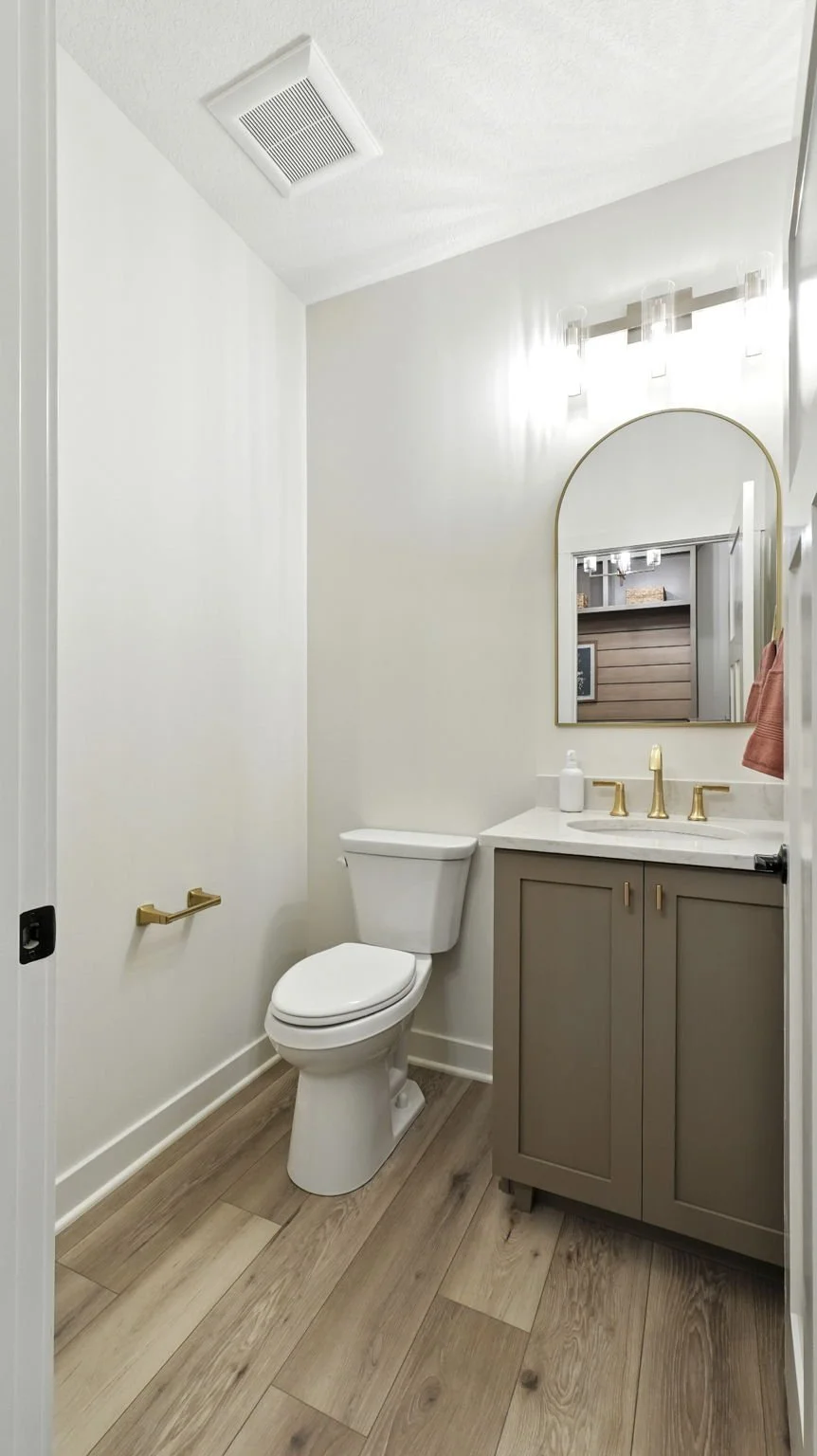 A small modern bathroom with a toilet, a vanity with gold fixtures, a mirror, a wall-mounted light fixture, and wood-look flooring.