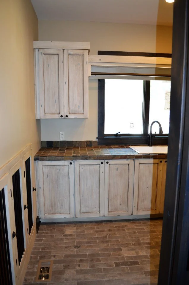 A small kitchen with whitewashed wooden cabinets, a tiled countertop, a large window above the sink, and a brown wood-floor pattern.