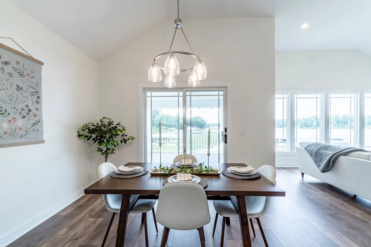 Dining room with wooden table, six white chairs, hanging light fixture, sliding glass door leading outside, and a living room with large windows and a white sofa with a grey blanket.