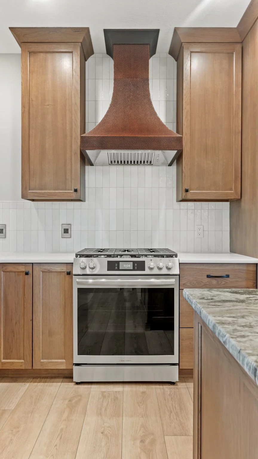 View of a modern kitchen with wood cabinets, a white tiled backsplash, a stainless steel stove, and a copper-colored range hood.