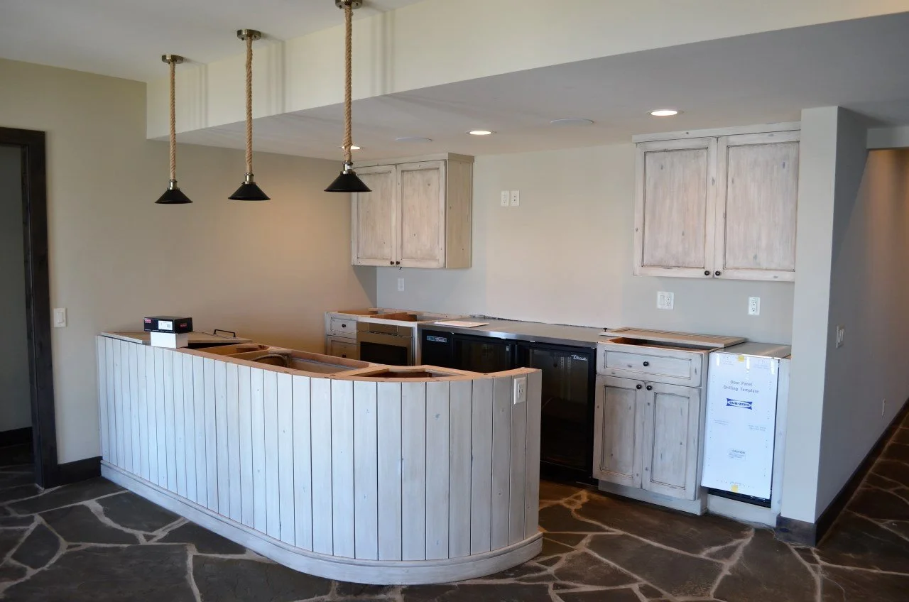 Unfinished kitchen with light wood cabinets, black appliances, and a curved wooden breakfast bar. The floor has a stone pattern, and there's a three pendant light hanging from the ceiling.