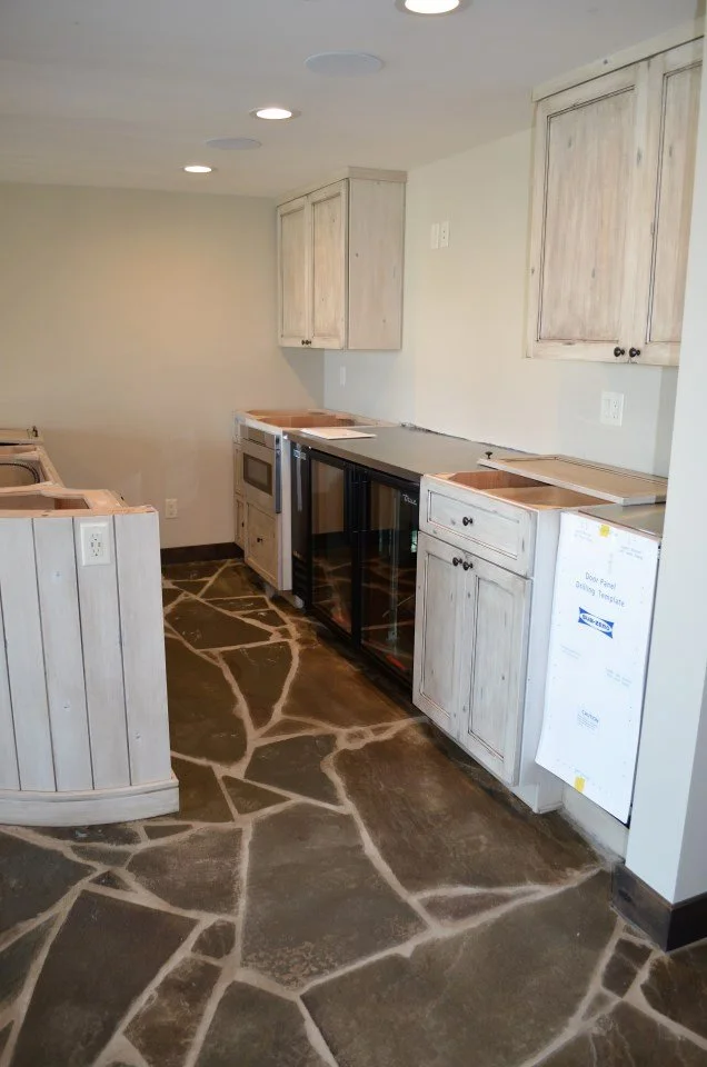 Kitchen area with unfinished cabinetry, a small refrigerator, and a stove. The floor has a patchwork stone pattern. Cabinets are light-colored wood with black knobs, and the wall is painted a light neutral color.