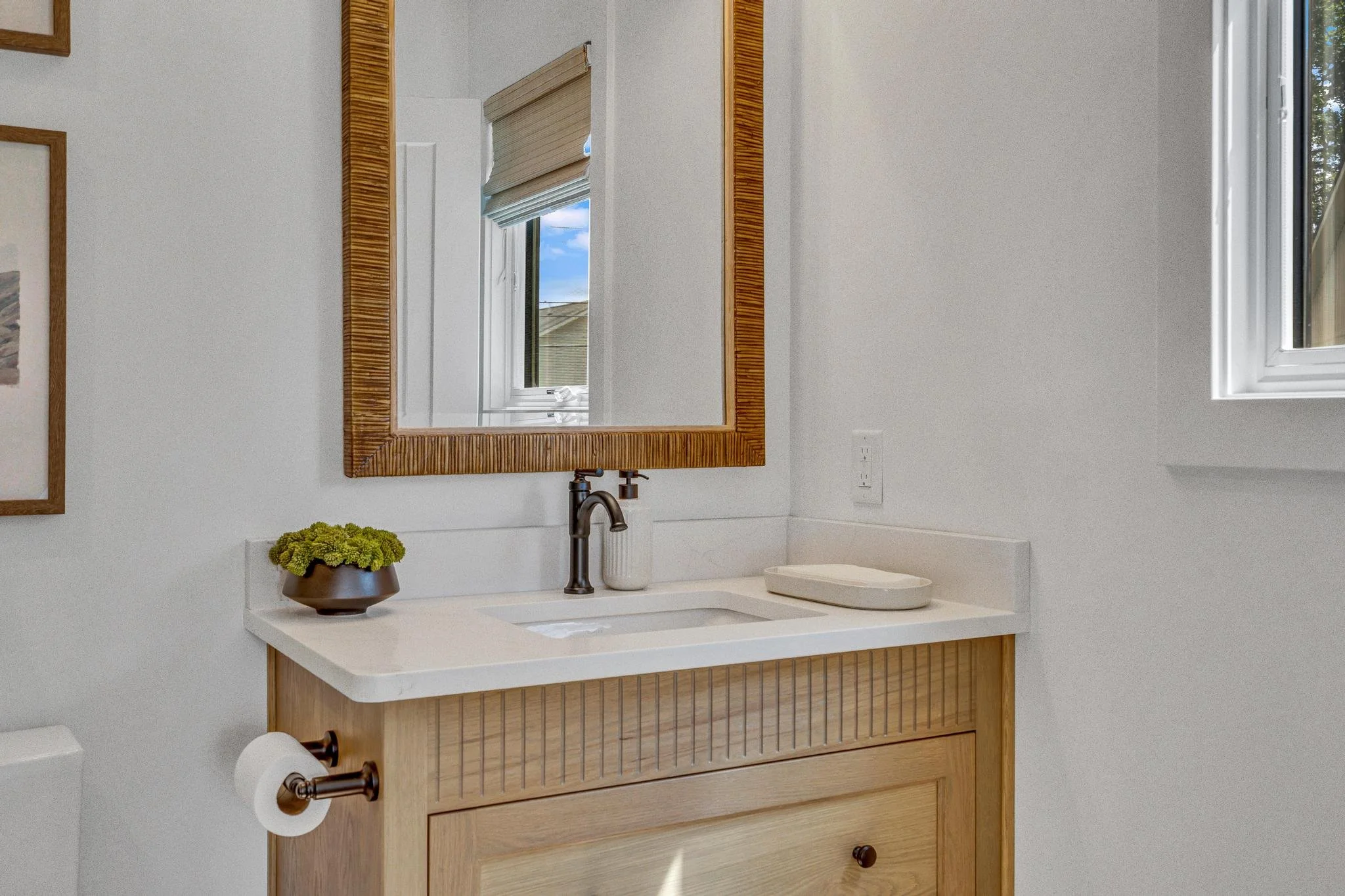A bathroom vanity with a white countertop, a small sink, a dark faucet, a mirror with a wooden frame, a small brown pot with green plants, a soap dish, a roll of toilet paper, white walls, and a window with a view of a house and blue sky.