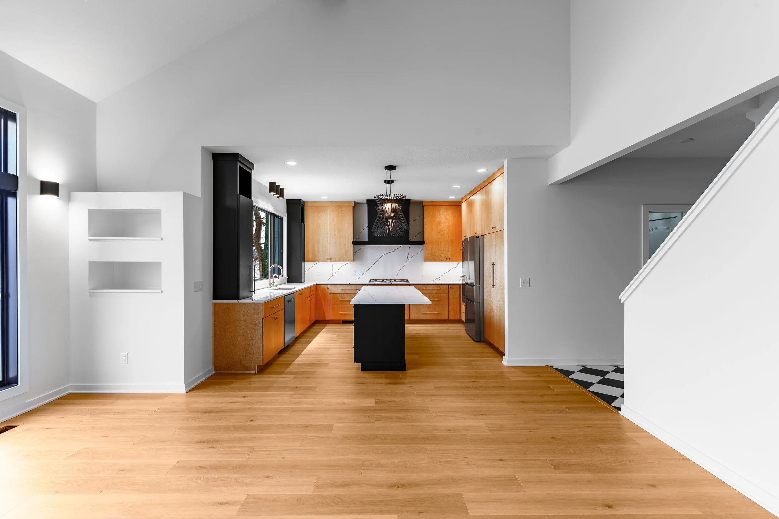 Modern kitchen with wooden cabinets, black accents, white marble backsplash, stainless steel appliances, a kitchen island with a black base and white countertop, and hardwood flooring.