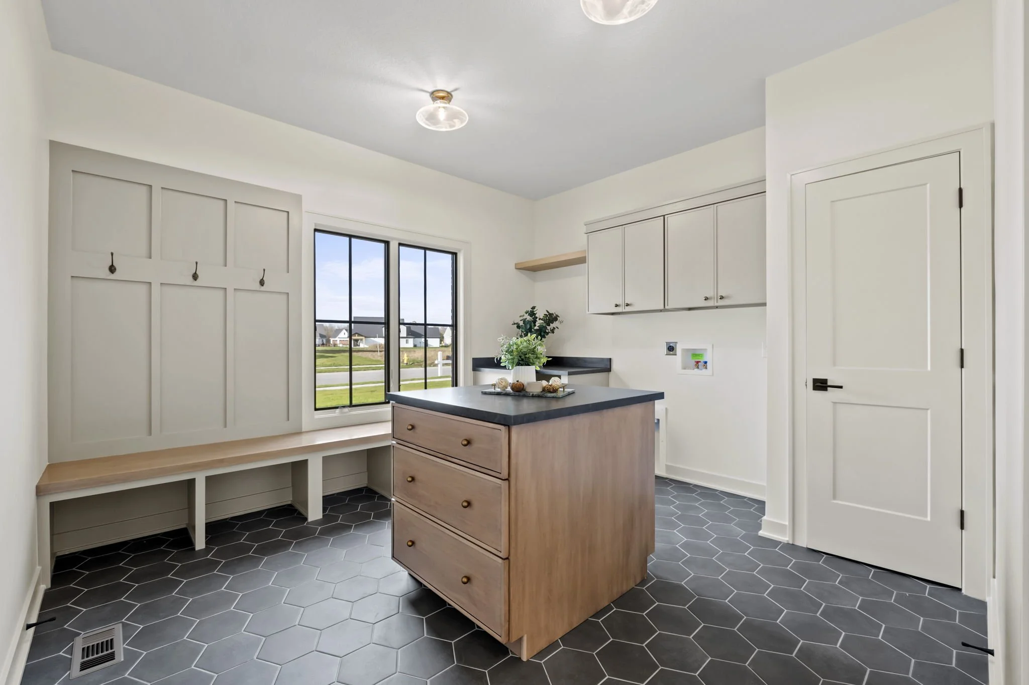 Empty kitchen with white cabinets, black countertops, a small island with drawers, hexagon gray floor tiles, a window overlooking a neighborhood, and a built-in bench with hooks on the wall.