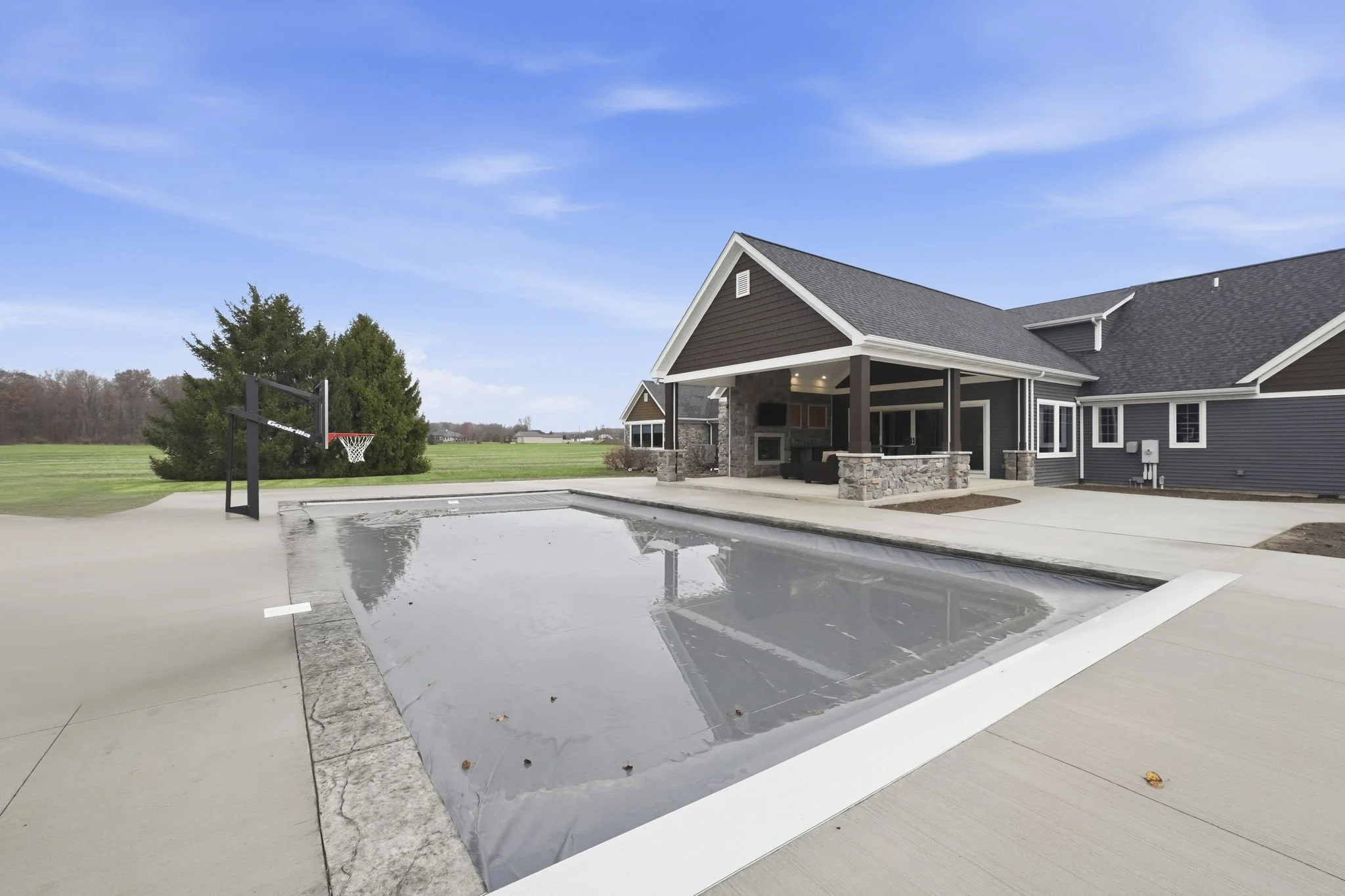 A backyard with a covered patio area, a small painted basketball hoop, and a closed-in in-ground pool covered with a safety cover, adjacent to a modern house with dark siding and a gray roof under a blue sky.