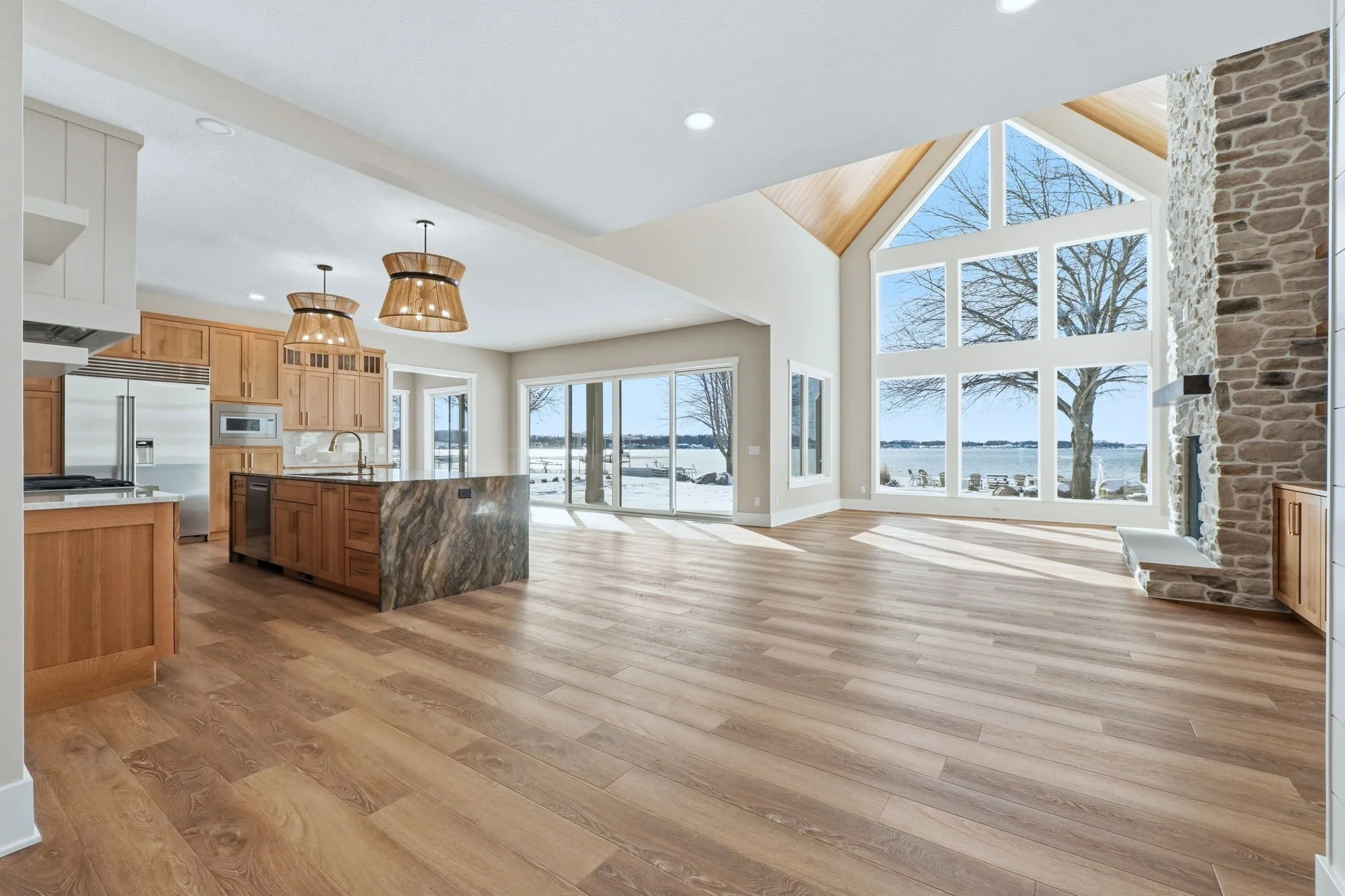 Modern kitchen and living area with large windows showcasing a snowy lakeside landscape, wooden floors, stone fireplace, and contemporary decor.