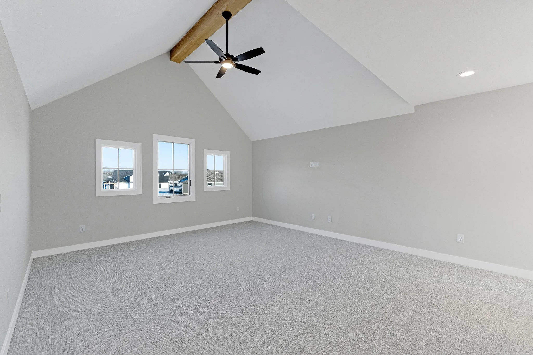 Empty room with light gray walls, white trim, three small windows, a ceiling fan with black blades and a wooden beam attached to the ceiling, and carpeted floor.
