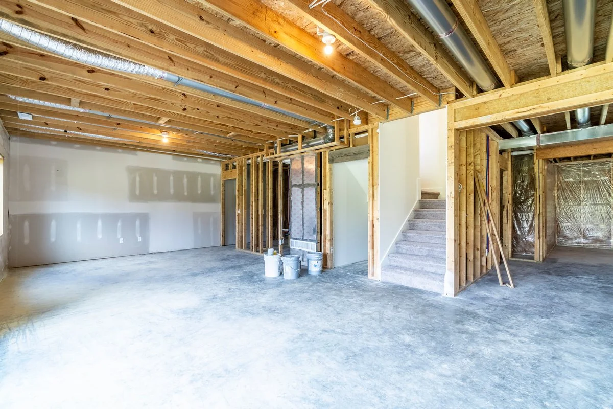 Interior of a house under construction with exposed wooden framing, ductwork, and unfinished flooring. There is a staircase with carpeted steps, and construction materials on the floor.