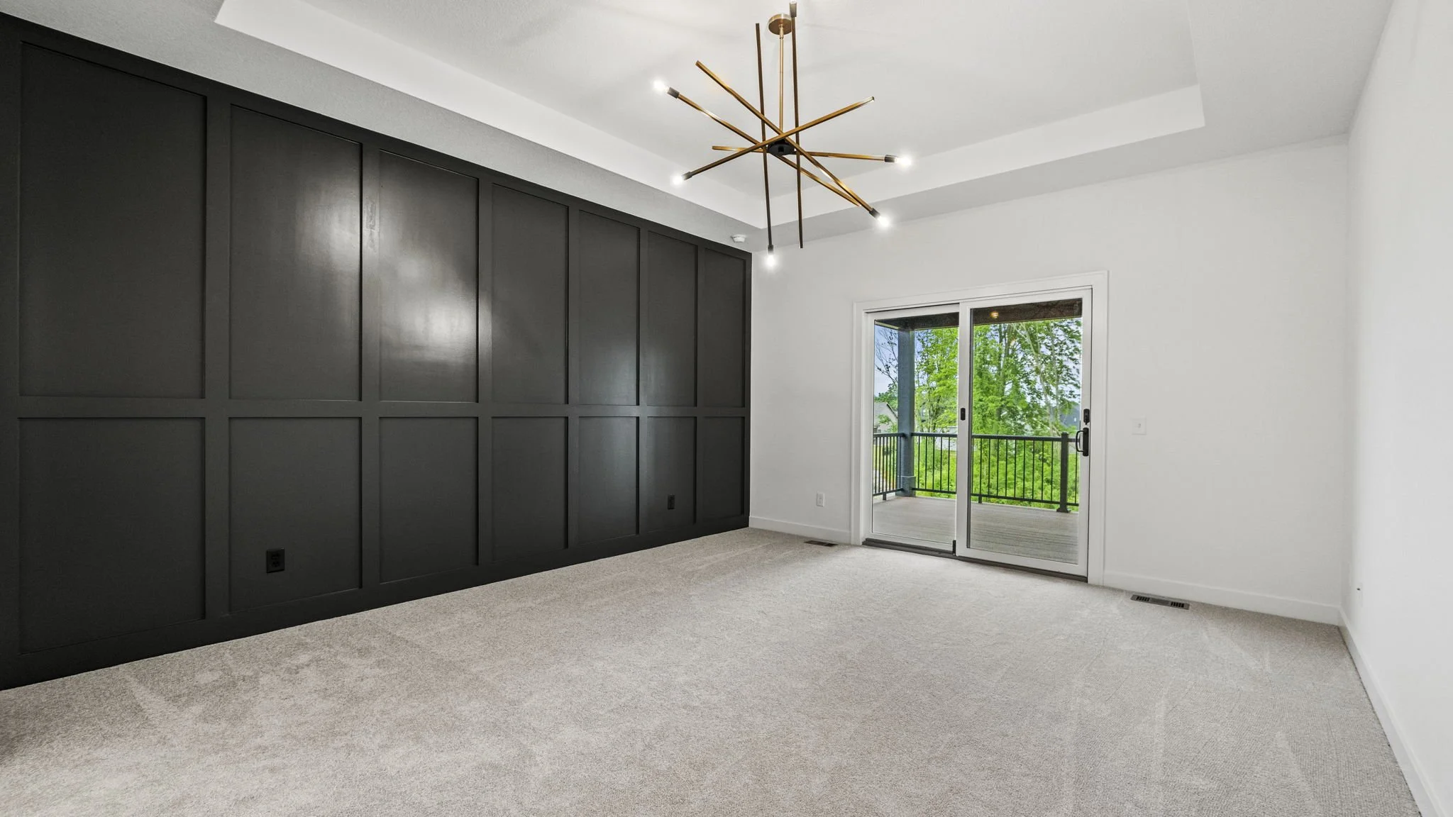 Empty room with dark paneled wall, beige carpet, white walls, a modern chandelier, and a sliding glass door leading to a balcony with trees outside.