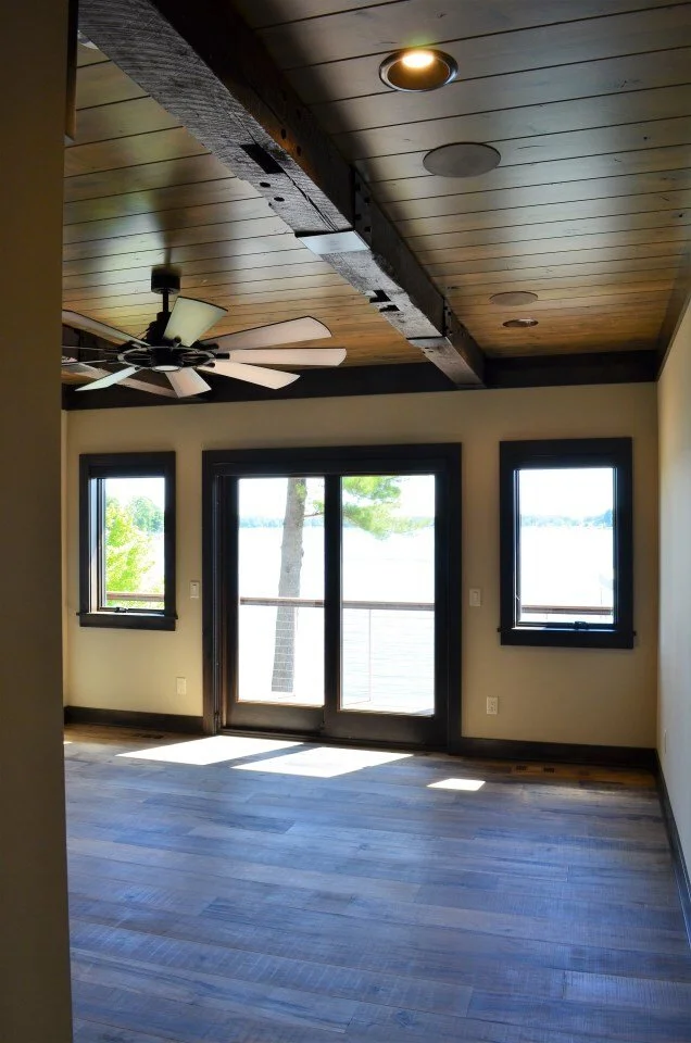 Empty room with wooden flooring, large glass door leading to a balcony overlooking water, and two smaller windows showing trees outside. Ceiling has wooden panels and a ceiling fan with lights.
