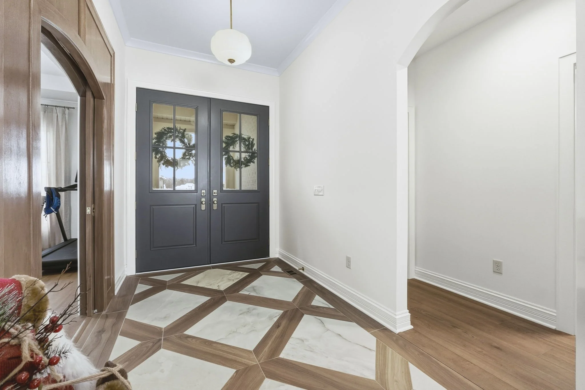 Interior view of a house entryway with a dark gray front door with glass panels, white walls, wood and marble patterned floor tiles, and a ceiling light fixture.