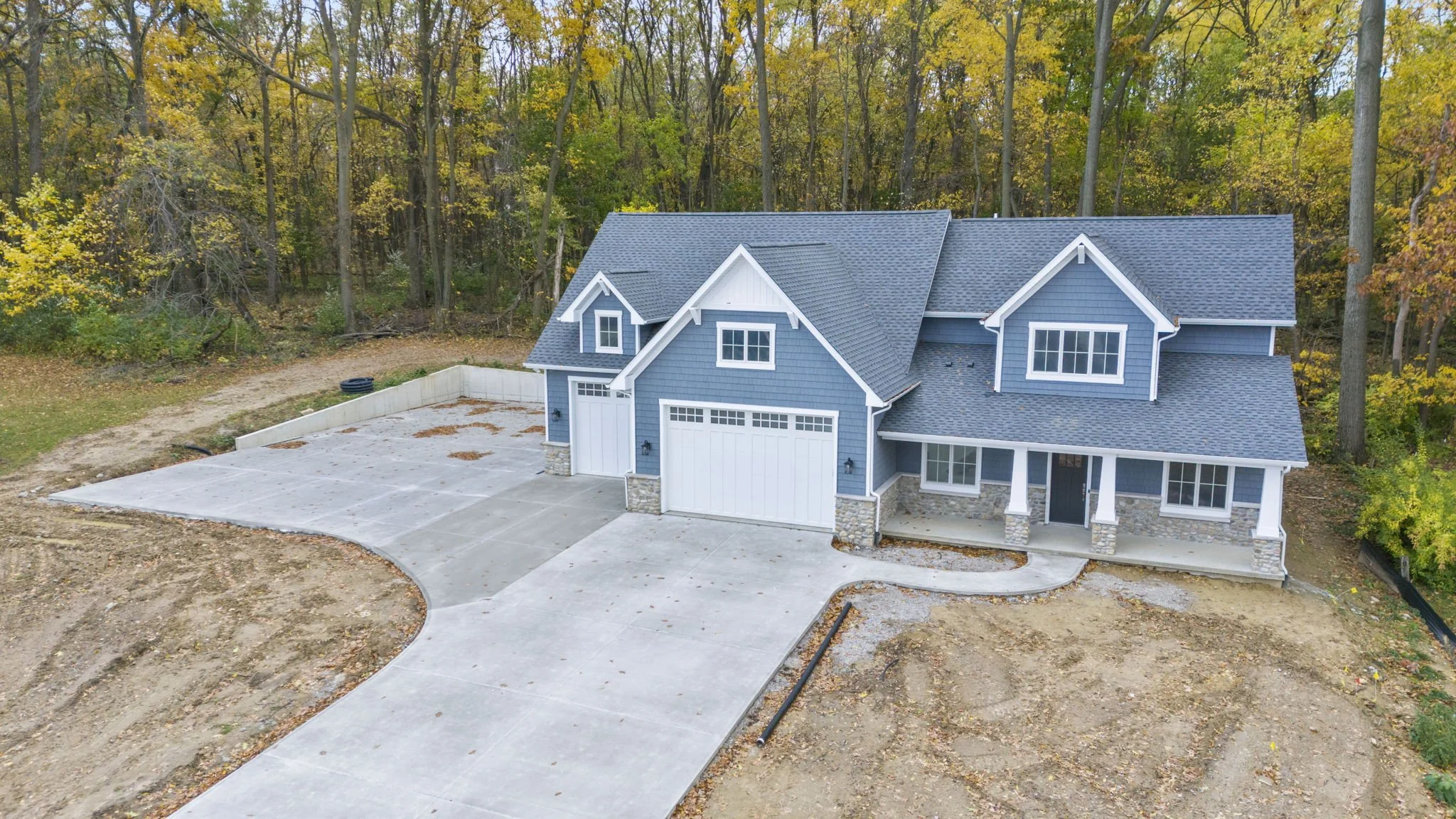 A newly built two-story house with blue siding, stone accents, and a large driveway, surrounded by fall trees.