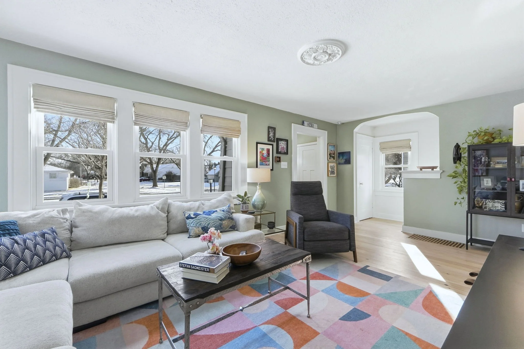 Bright living room with large windows, white couch with patterned pillows, black armchair, colorful geometric rug, and a coffee table with books and a bowl.