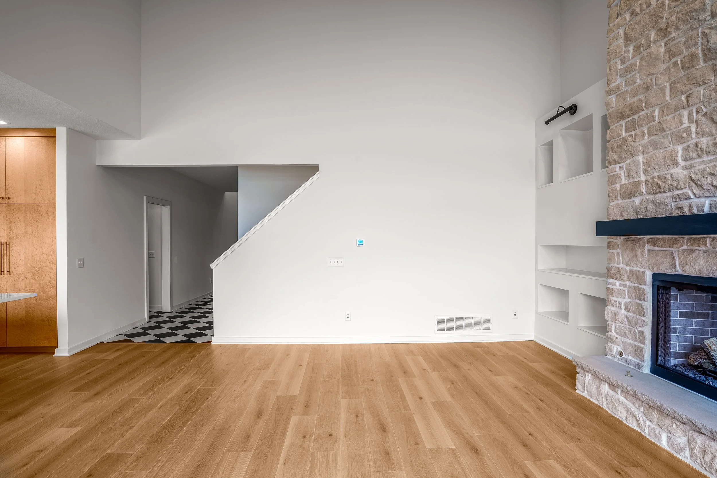 Empty living room with white walls, wood flooring, stone fireplace, and built-in shelves, leading to hallway with black and white checkered tile floor.