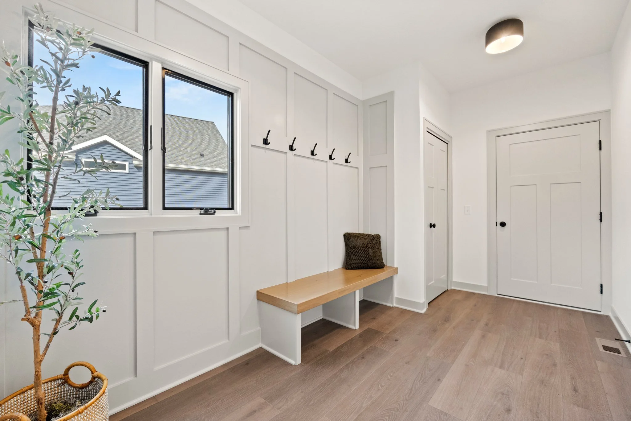 A white mudroom with a window, a wooden bench with a dark cushion, black hooks on the wall, and a potted plant in the corner.