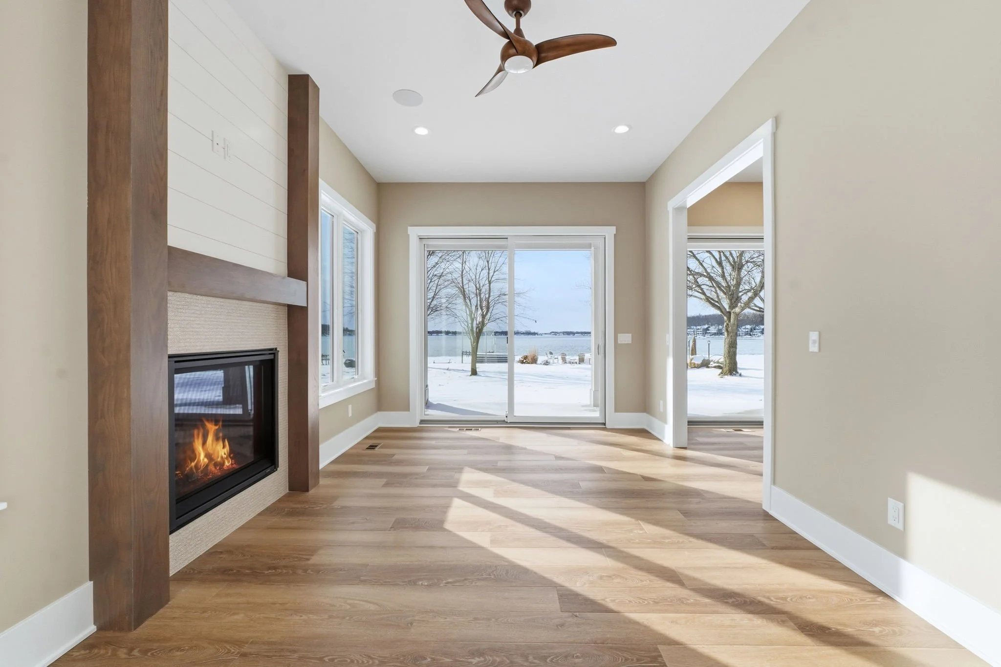 Empty living room with a fireplace, hardwood floors, large windows, sliding glass door leading outside to snowy yard, and a ceiling fan.