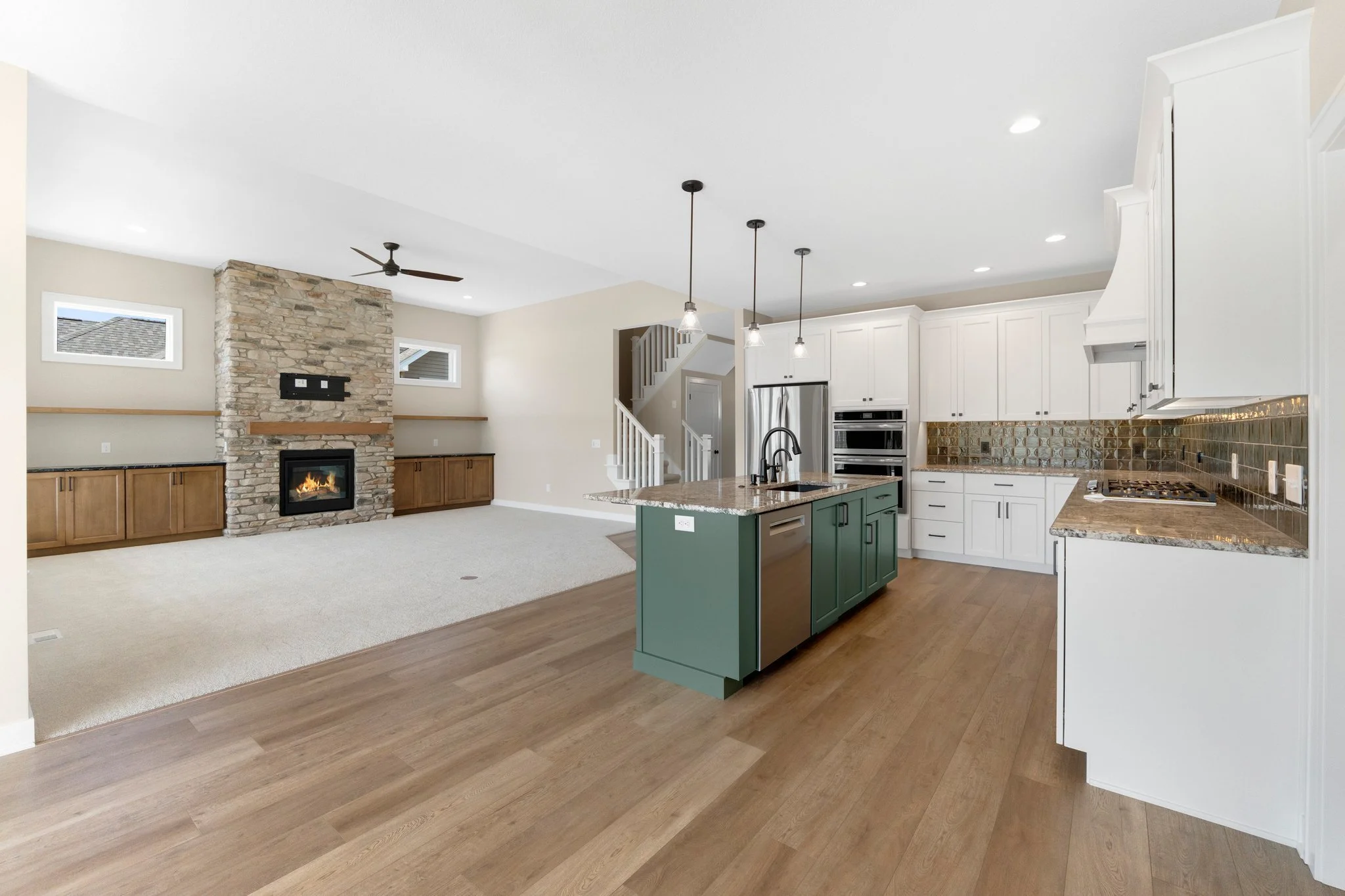 Open-concept living room and kitchen with a stone fireplace, wooden built-in cabinets, white cabinets with granite countertops, a green kitchen island with a granite top, stainless steel appliances, and hardwood flooring.