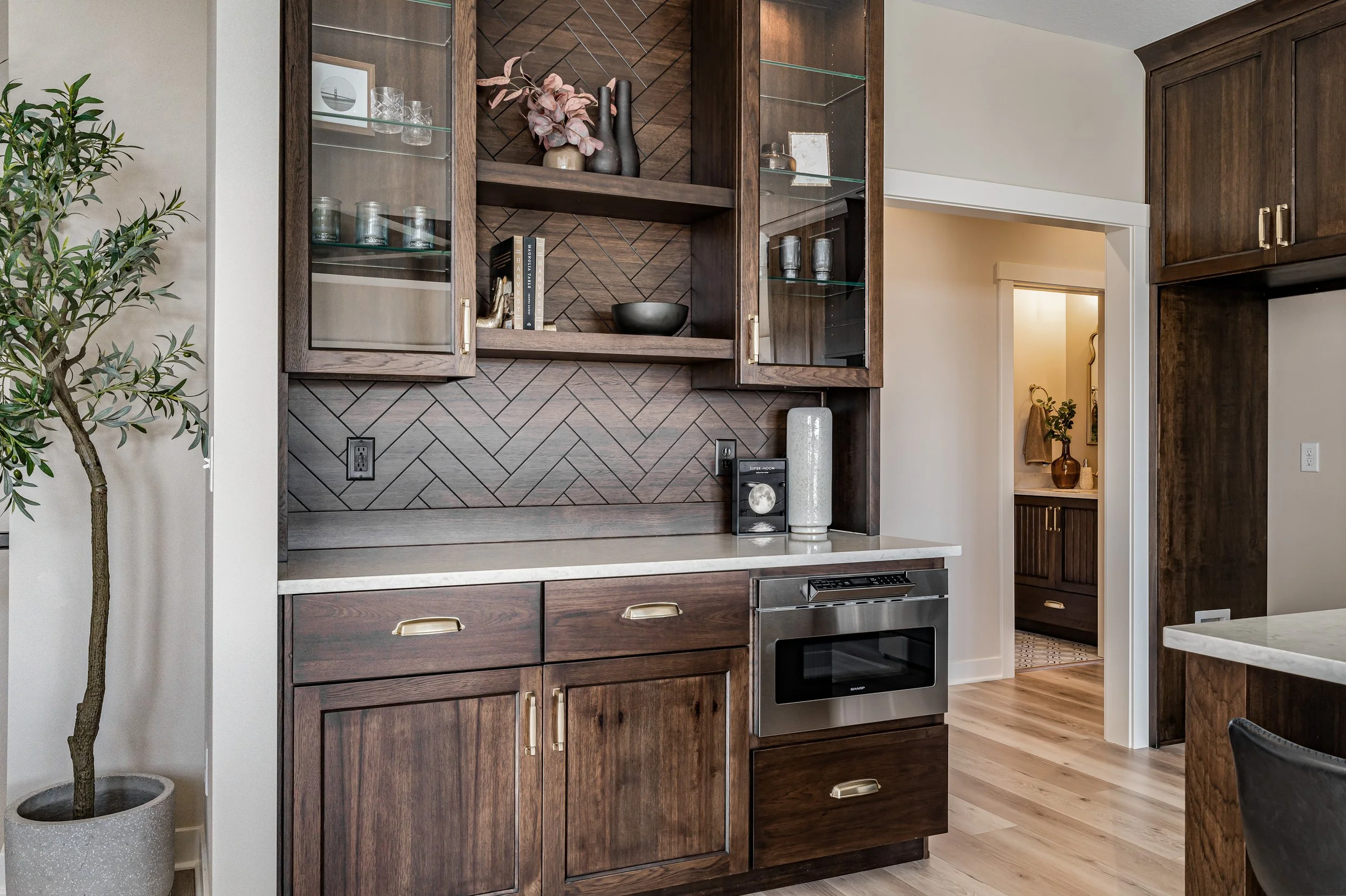 A kitchen corner with dark wooden cabinetry, a built-in oven, open shelves, and a herringbone patterned backsplash. There is a potted plant to the left and decorative items on the shelves.