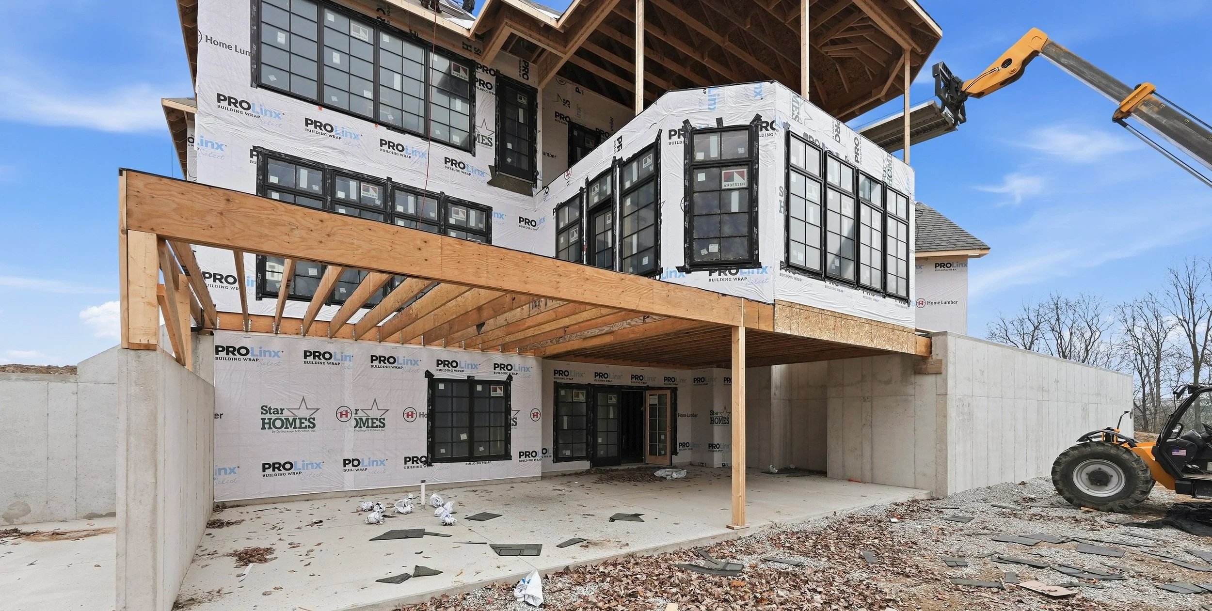 A multi-story house under construction with wrapped exterior insulation and black window frames. The house has a wooden deck area on the second floor, partially supported by the ground level. Construction equipment, including a crane arm, is visible in the scene, along with a concrete wall and a construction vehicle on the right. The sky is clear and blue.