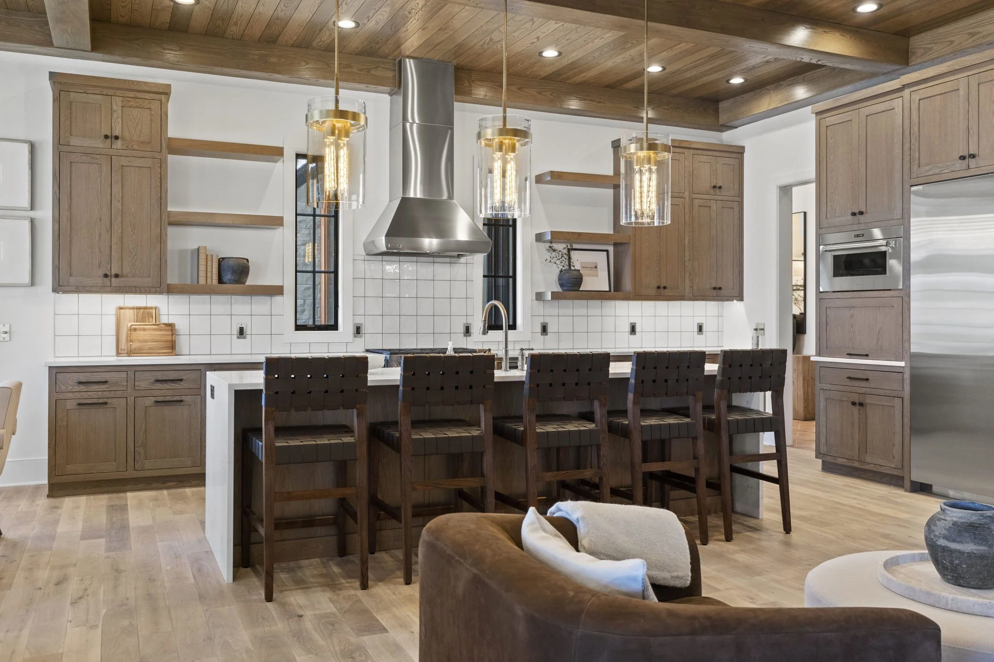 Modern kitchen with wooden cabinets, a white tiled backsplash, a stainless steel range hood, and designed pendant lighting over a kitchen island with four dark brown cast-iron barstools. There's a window between the cabinets, and a portion of a livin