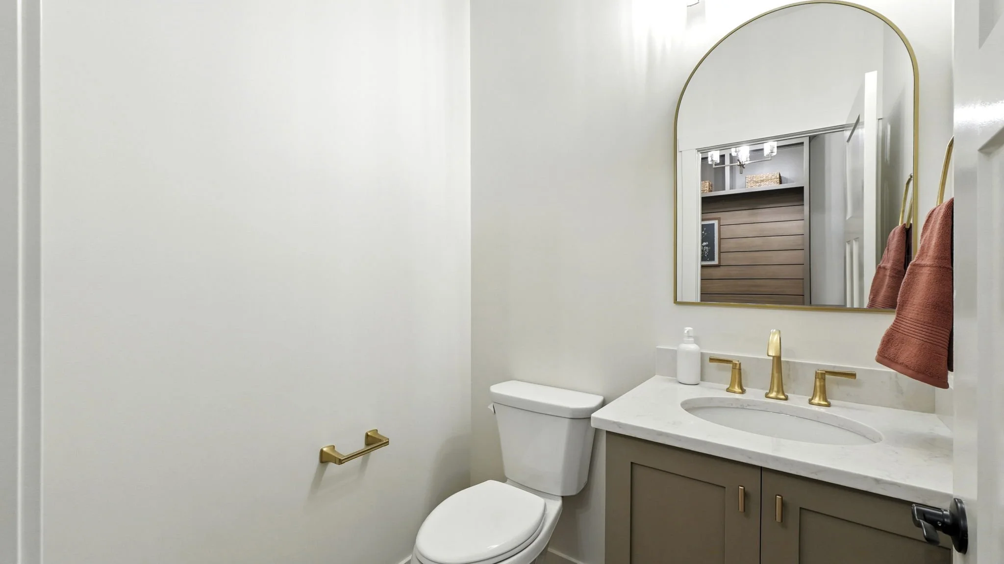 A small, modern bathroom with white walls, a toilet, a beige vanity with a marble countertop, a gold faucet, and a large arched mirror. Red towels hang on a gold towel rack, and a soap dispenser is on the sink.