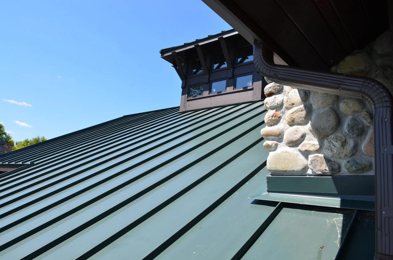 Close-up of a green metal roof, a stone chimney, and a dark brown wooden dormer window under a clear blue sky.
