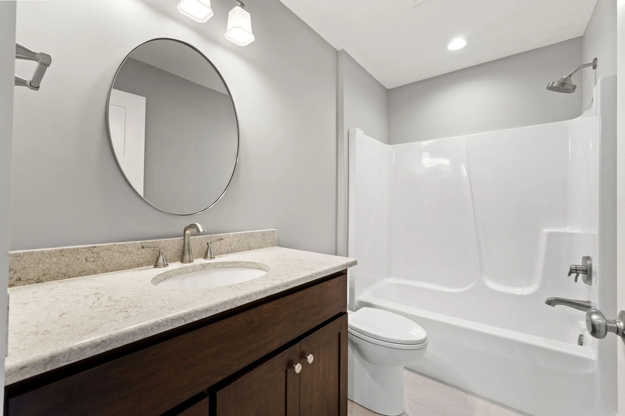 A bathroom with a granite countertop vanity with a round mirror above, a single sink with a silver faucet, a toilet, and a white bathtub with a showerhead, light gray walls, and ceiling lights.