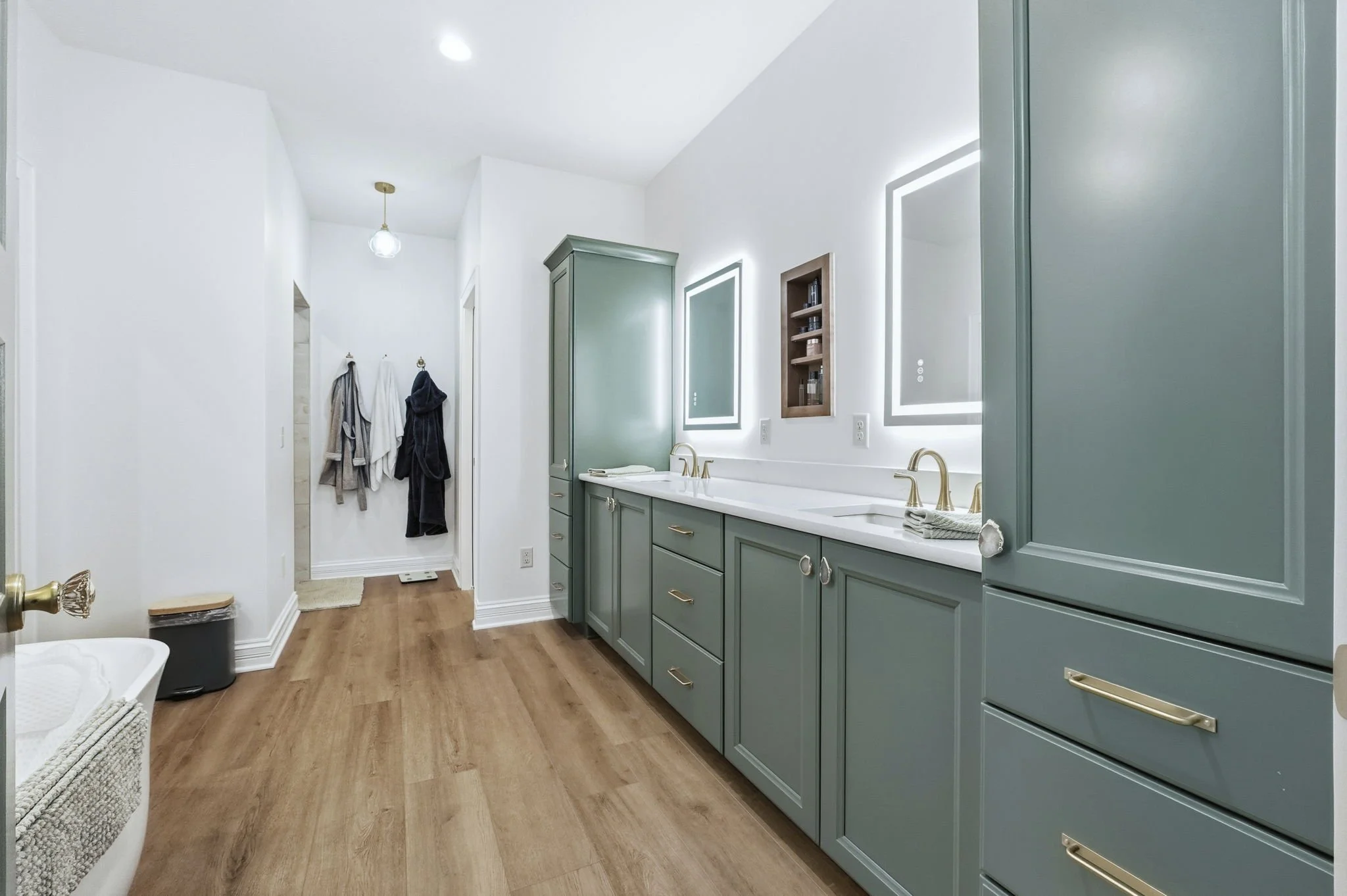 Modern bathroom with green cabinetry, double sinks, illuminated mirrors, and wooden floors.