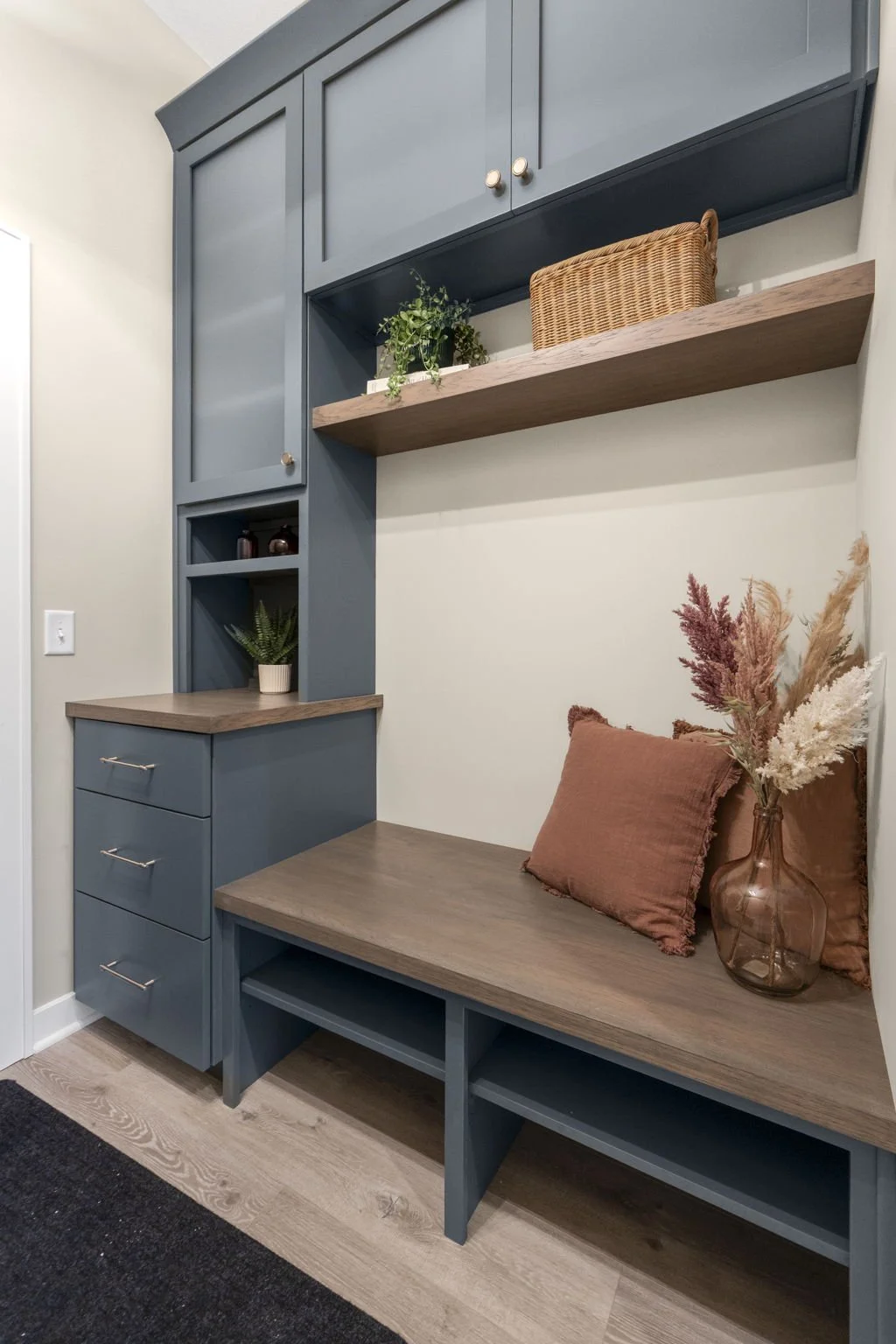 A built-in blue and wood bench with a cushioned seat, pillows, and decorative items, including a glass vase with dried flowers, situated under a large open shelf and cabinets in a modern interior.