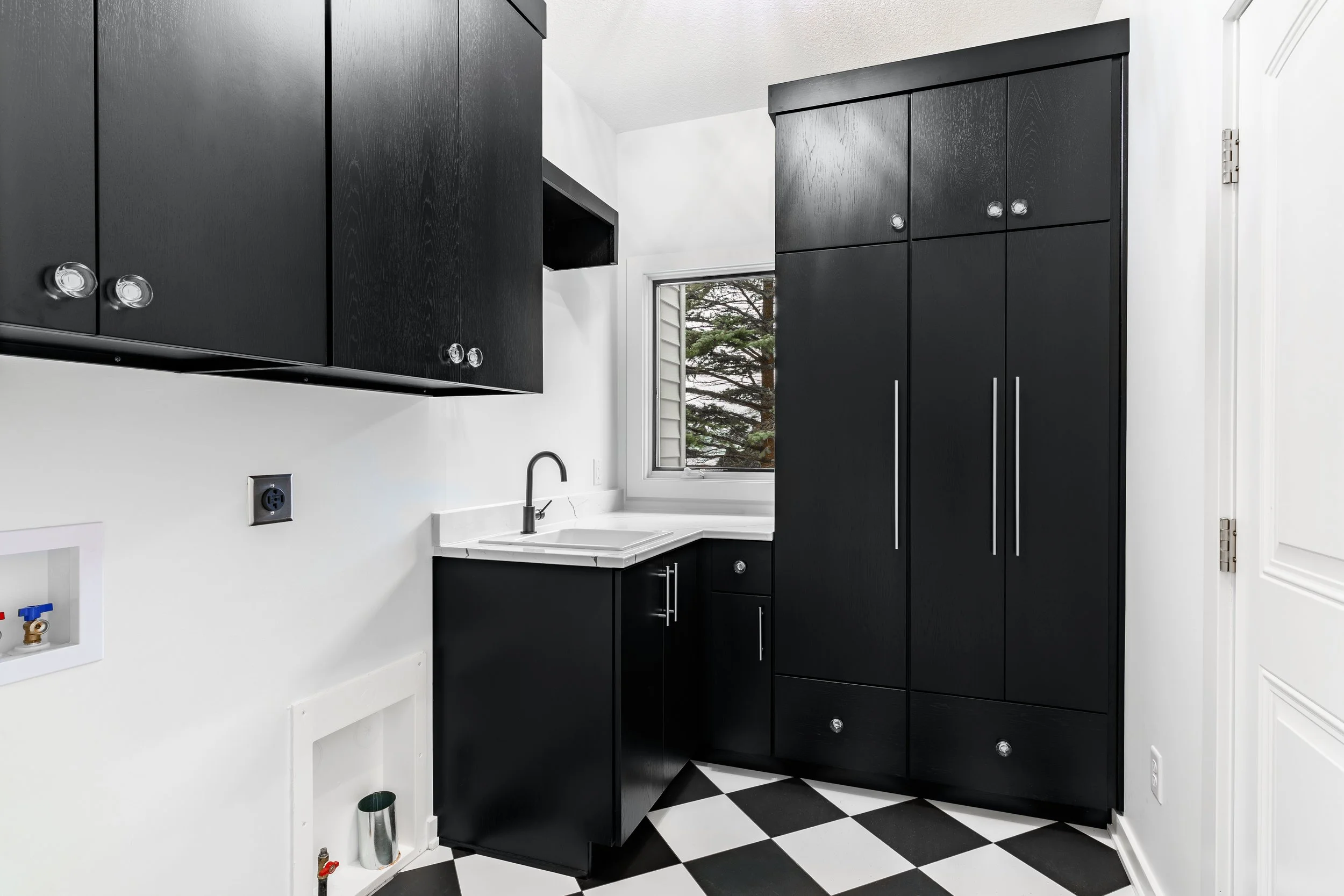 Kitchen with black cabinets, white countertops, black and white checkered floor, window with trees outside, black cabinets with silver handles, small sink, and a white door.