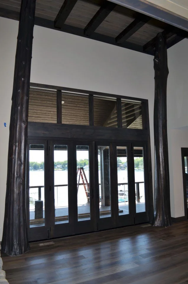 Interior of a room with large black framed glass doors opening to an outdoor deck overlooking water, with trees in the distance, and support beams inside.