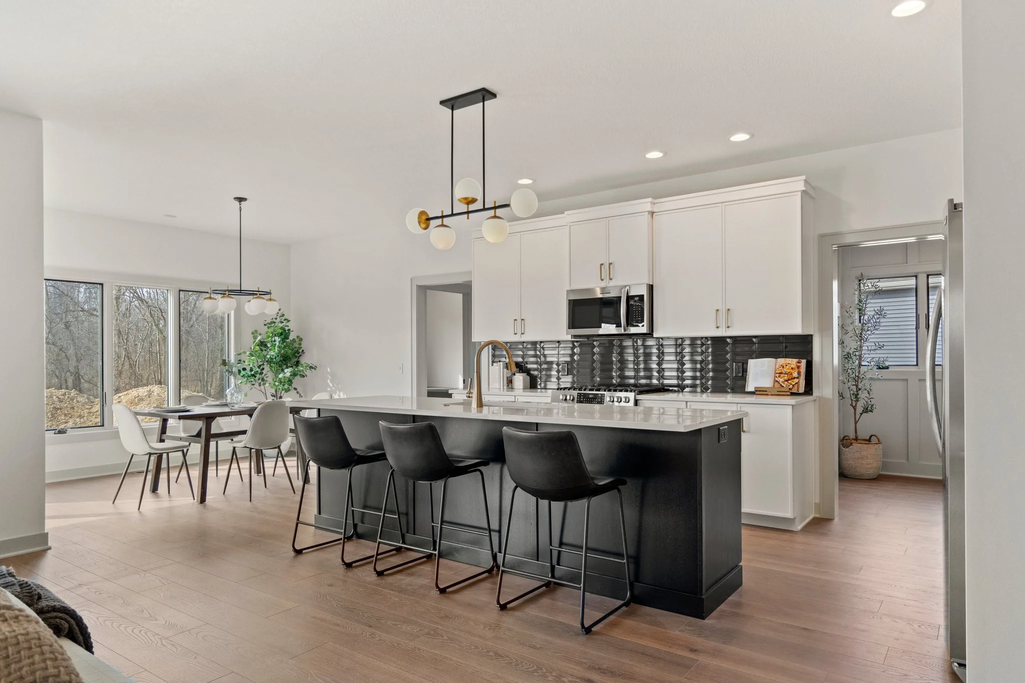 Modern kitchen with white cabinets, black and white patterned backsplash, black kitchen island with four barstools, and a dining area with large windows and a potted plant.