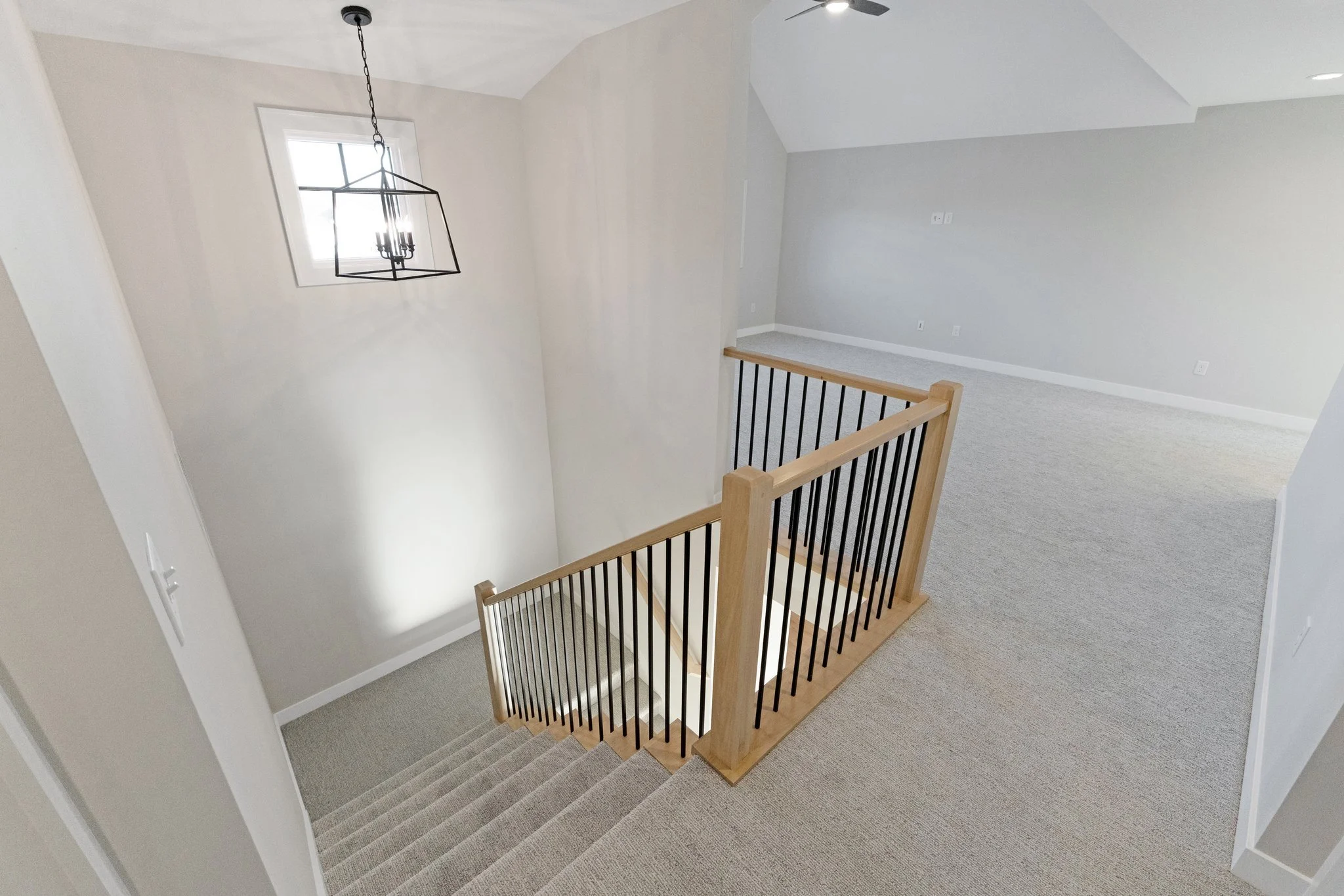 Interior view of a modern, empty house staircase area with beige carpet, a wooden and metal staircase railing, a wall-mounted light fixture, and a window with natural light.