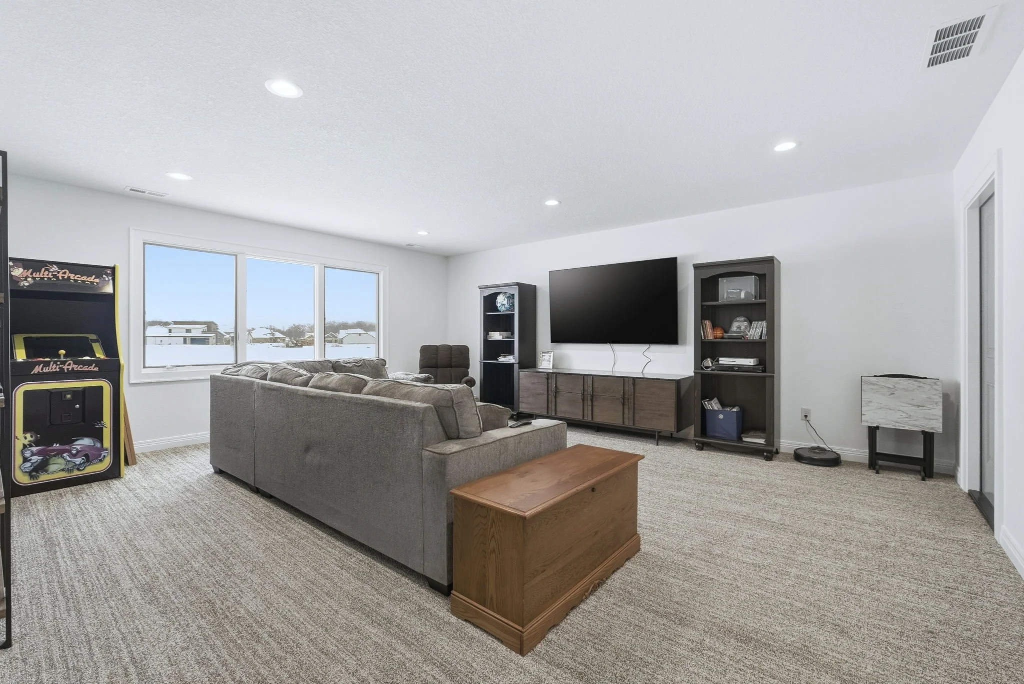Living room with beige sofa, wooden coffee table, large window with snow outside, TV on wall, arcade game, bookshelf, and carpeted floor.