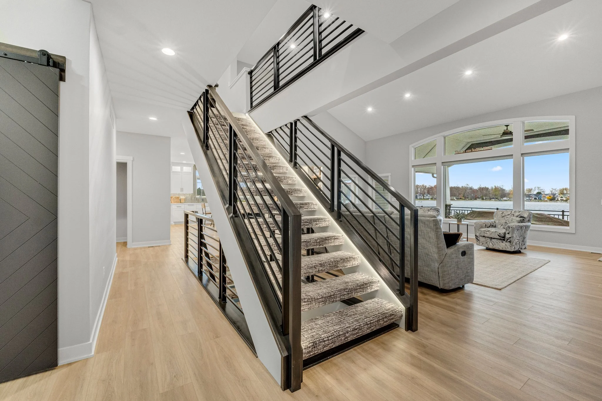 Empty living room with staircase, large window showing lake view, light wood flooring, and furniture