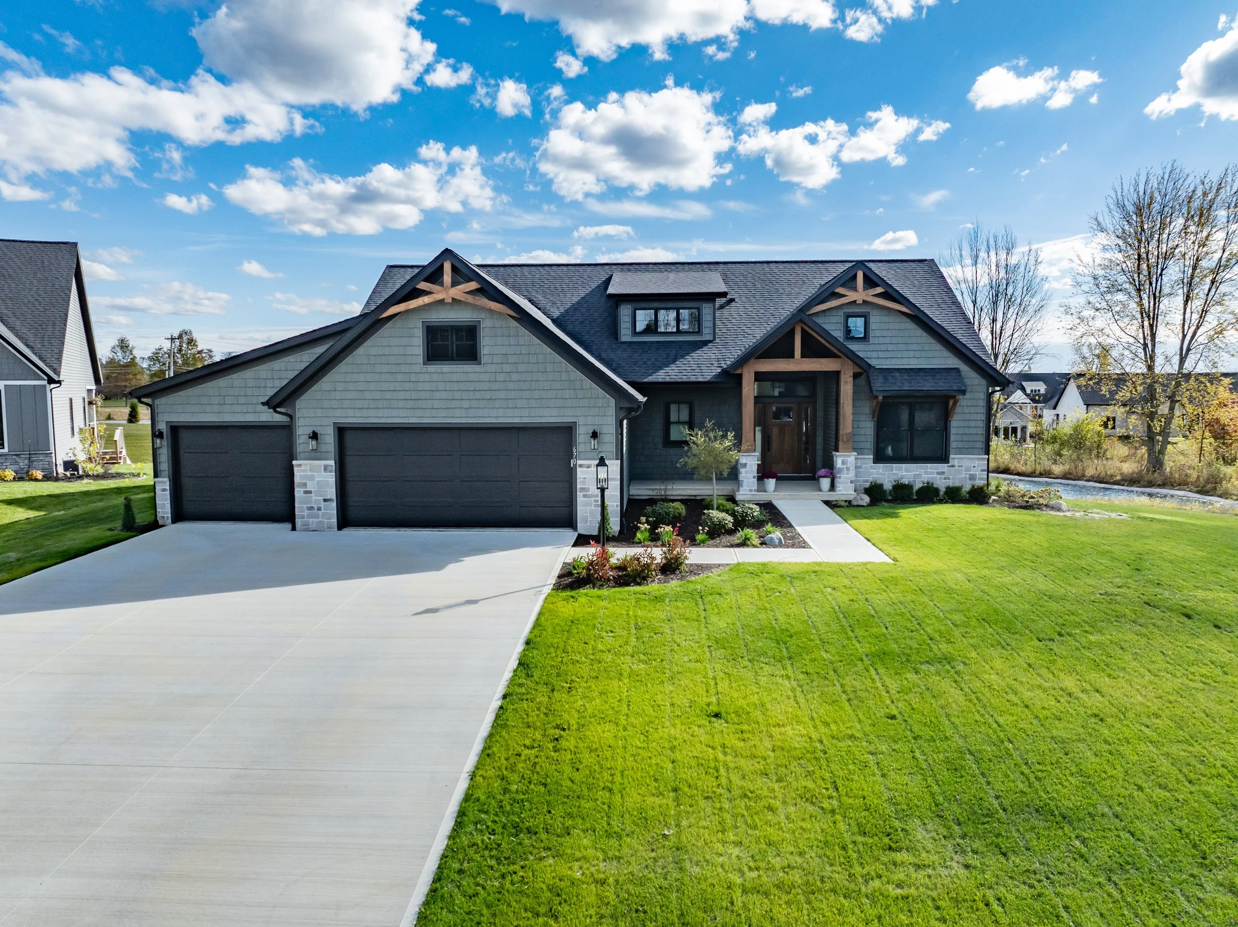 A modern house with a dark gray roof, gray siding, stone accents, and a wooden front door. The house has a two-car garage, a manicured green lawn, and a pathway leading to the front entrance under a blue sky with clouds.