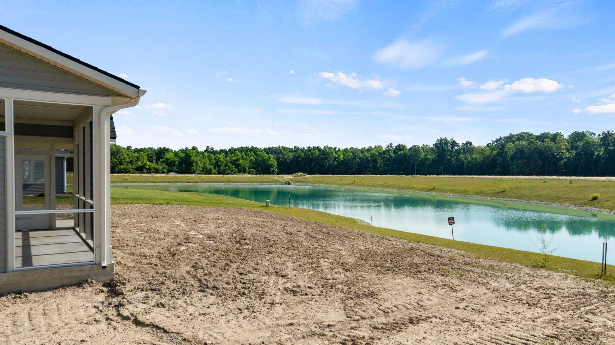 A house with a screened porch overlooking a pond and grassy landscape, with a wooded area and blue sky in the background.