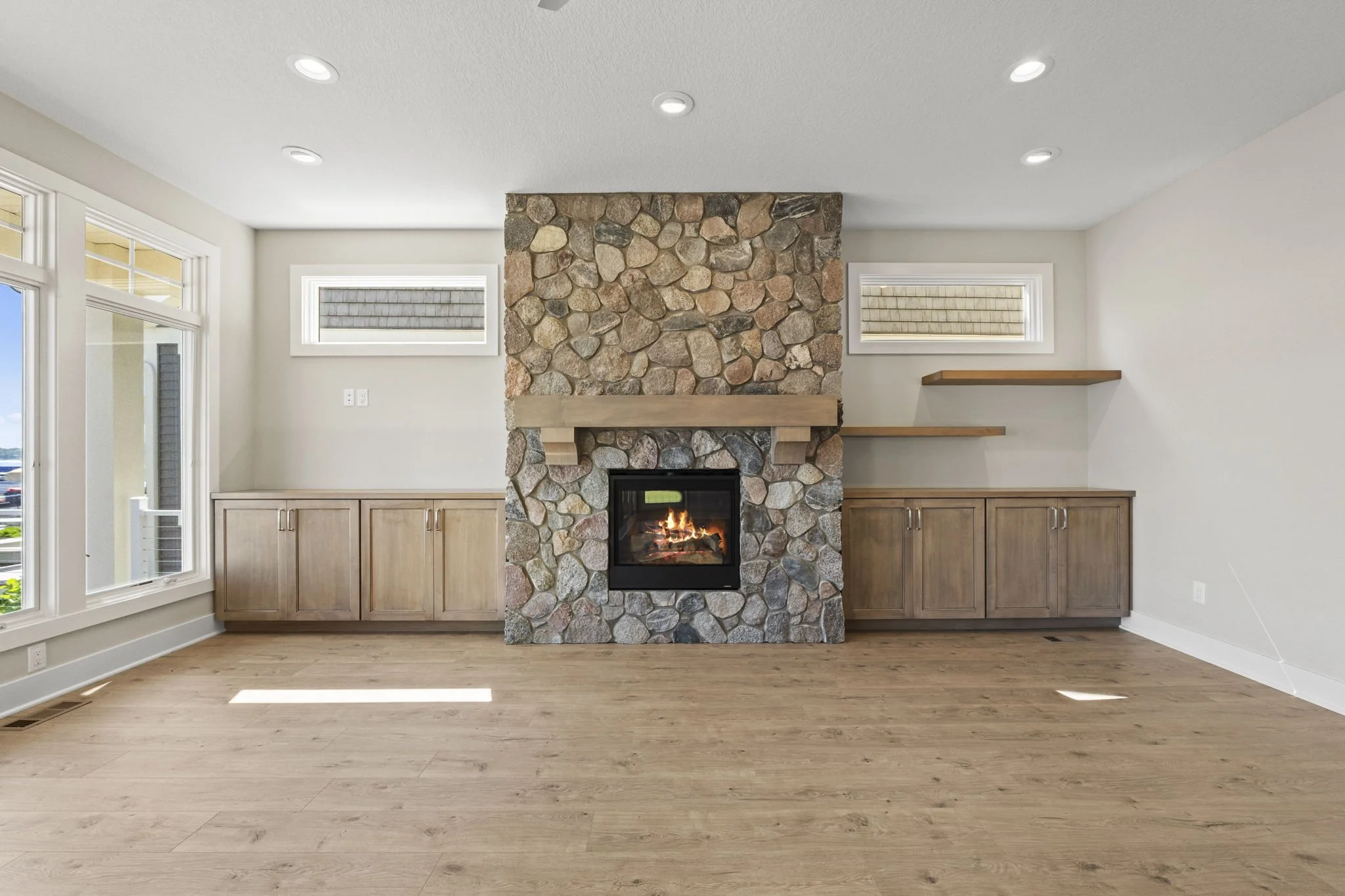 Empty living room with a stone fireplace, wooden shelves, and hardwood floors, illuminated by natural light from large windows.