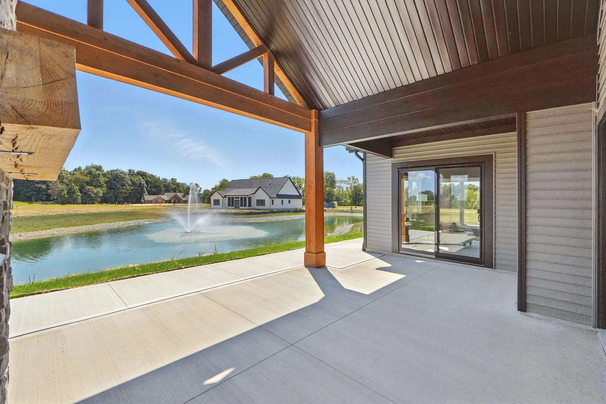 Backyard patio with a view of a pond, fountain, and neighboring houses, partially shaded by a wooden pergola.