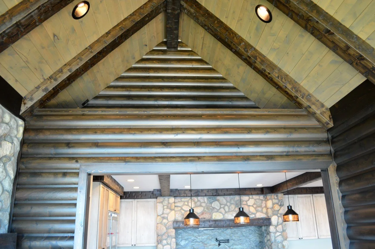 Interior view of a log cabin with a high, triangular ceiling, exposed wooden beams, and rustic stone fireplace with hanging pendant lights.