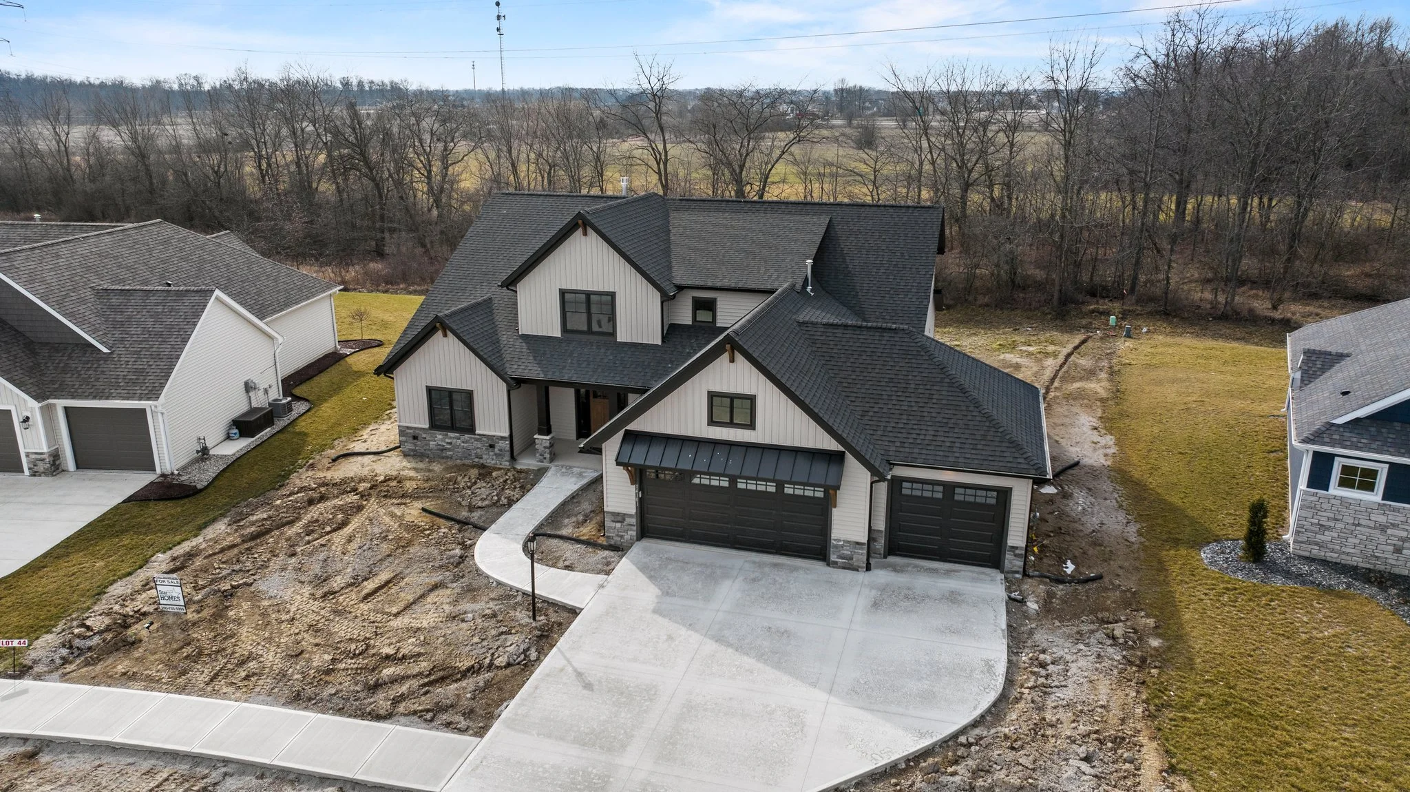 A large modern two-story house with a black shingle roof, white vertical siding, stone accents, and an attached black garage. The front yard has a concrete driveway and sidewalk, with nearby neighboring houses. In the background, there are bare trees