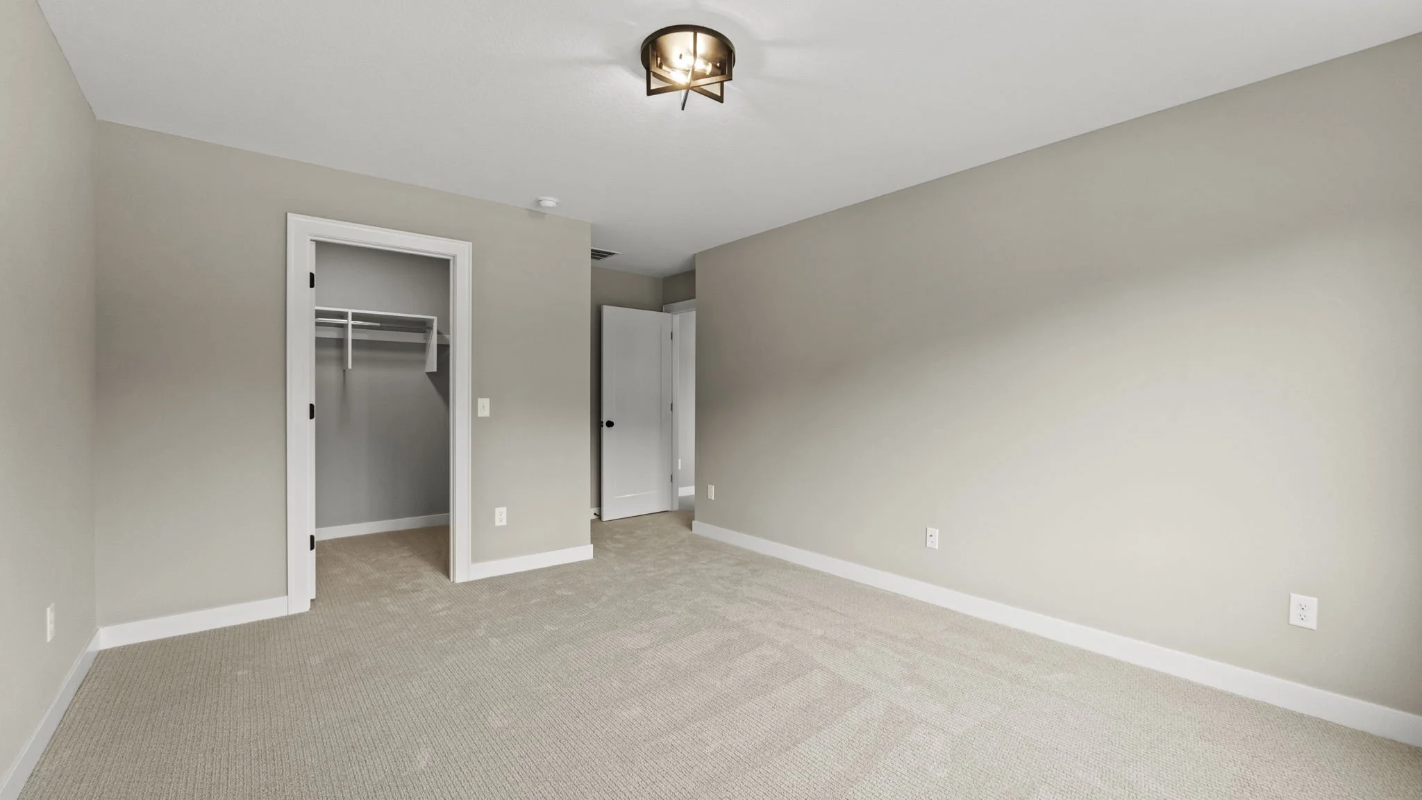 Empty beige carpeted bedroom with a closet, a white door, and a ceiling light fixture.