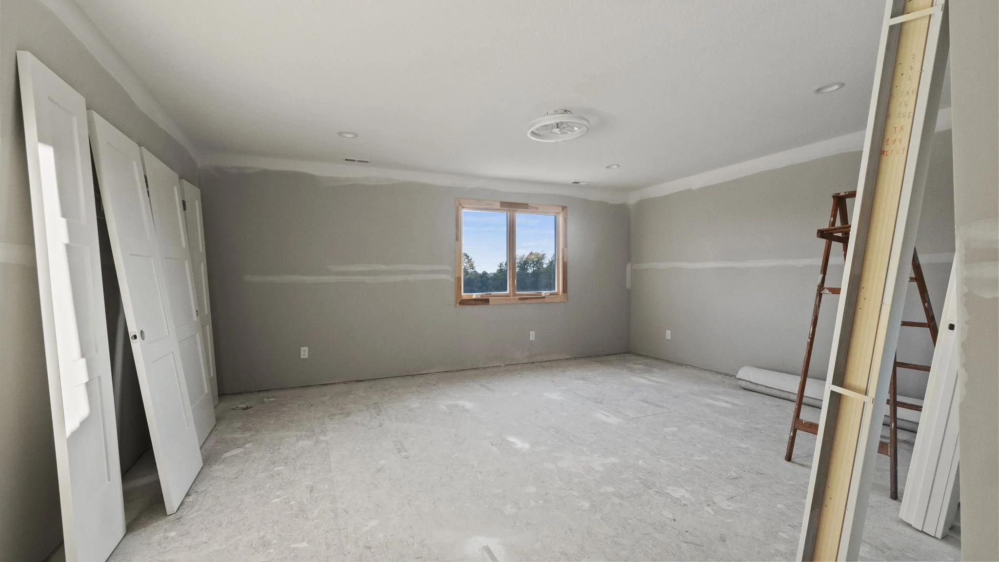 Empty room under construction with drywall and a window, with doors leaning against the wall and a ladder in the corner.