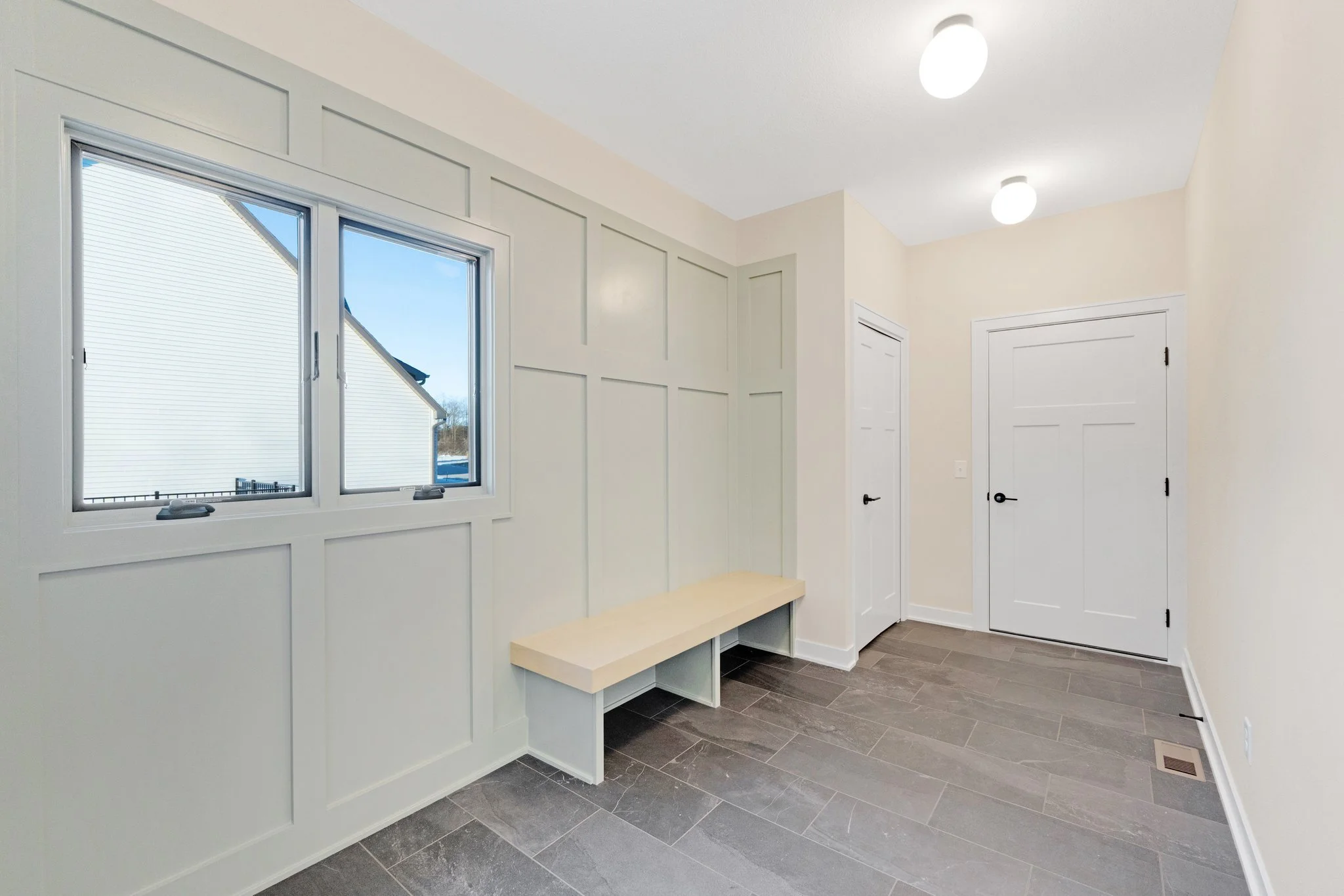 Empty room with a window on the left side and two closed white doors on the right, beige walls, a bench under the window, and ceiling lights.