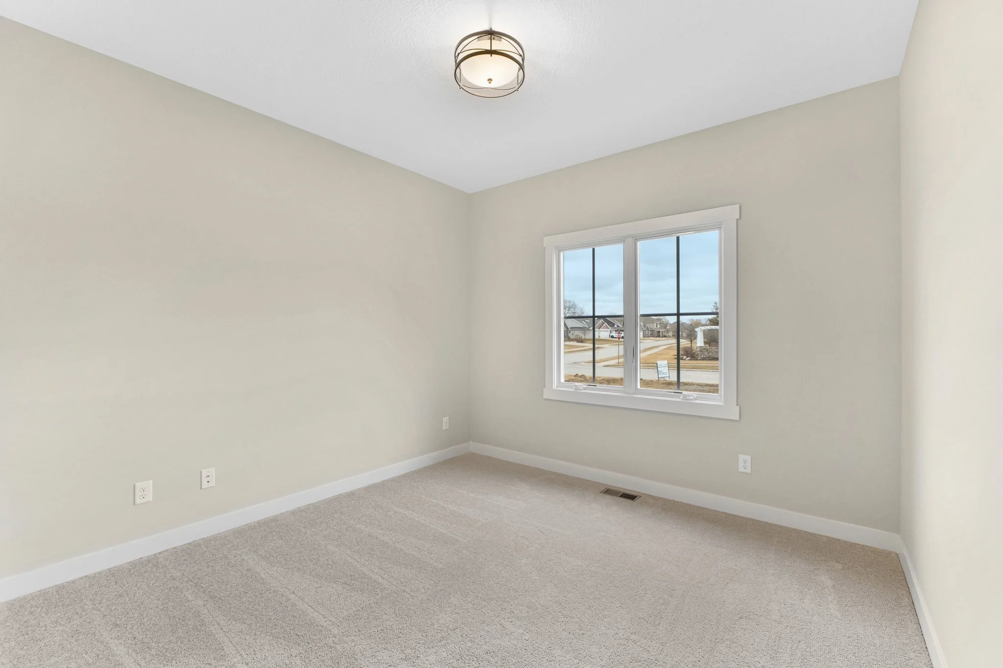 Empty room with beige carpet, white walls, a window with a view of a neighborhood street, and a ceiling light fixture.
